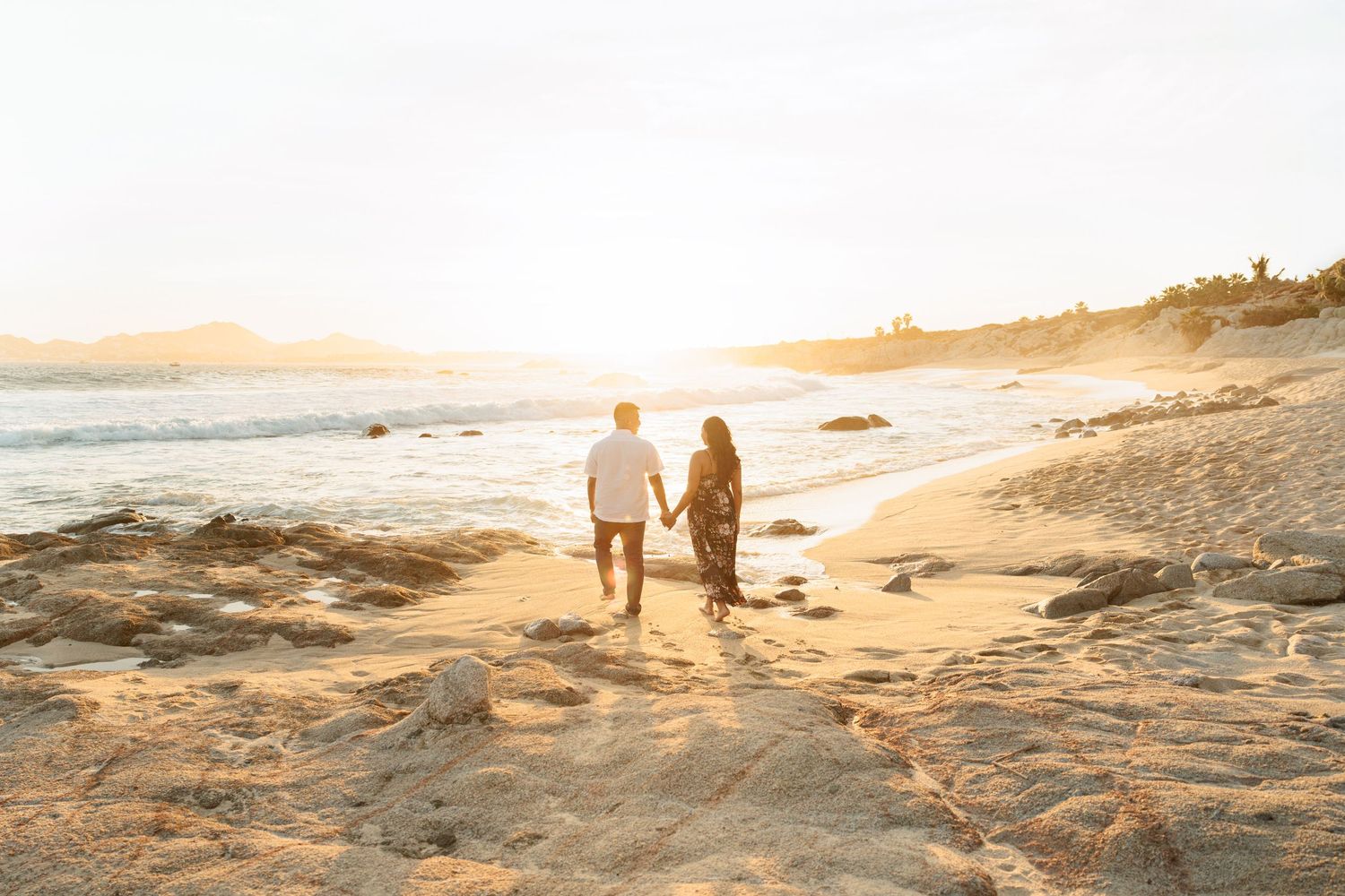 Couple walking hand in hand along a golden sandy beach toward a glowing sunset.