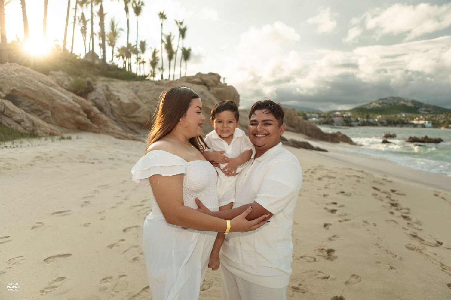 Happy family of three in white outfits posing at sunset on a palm tree-lined tropical beach.