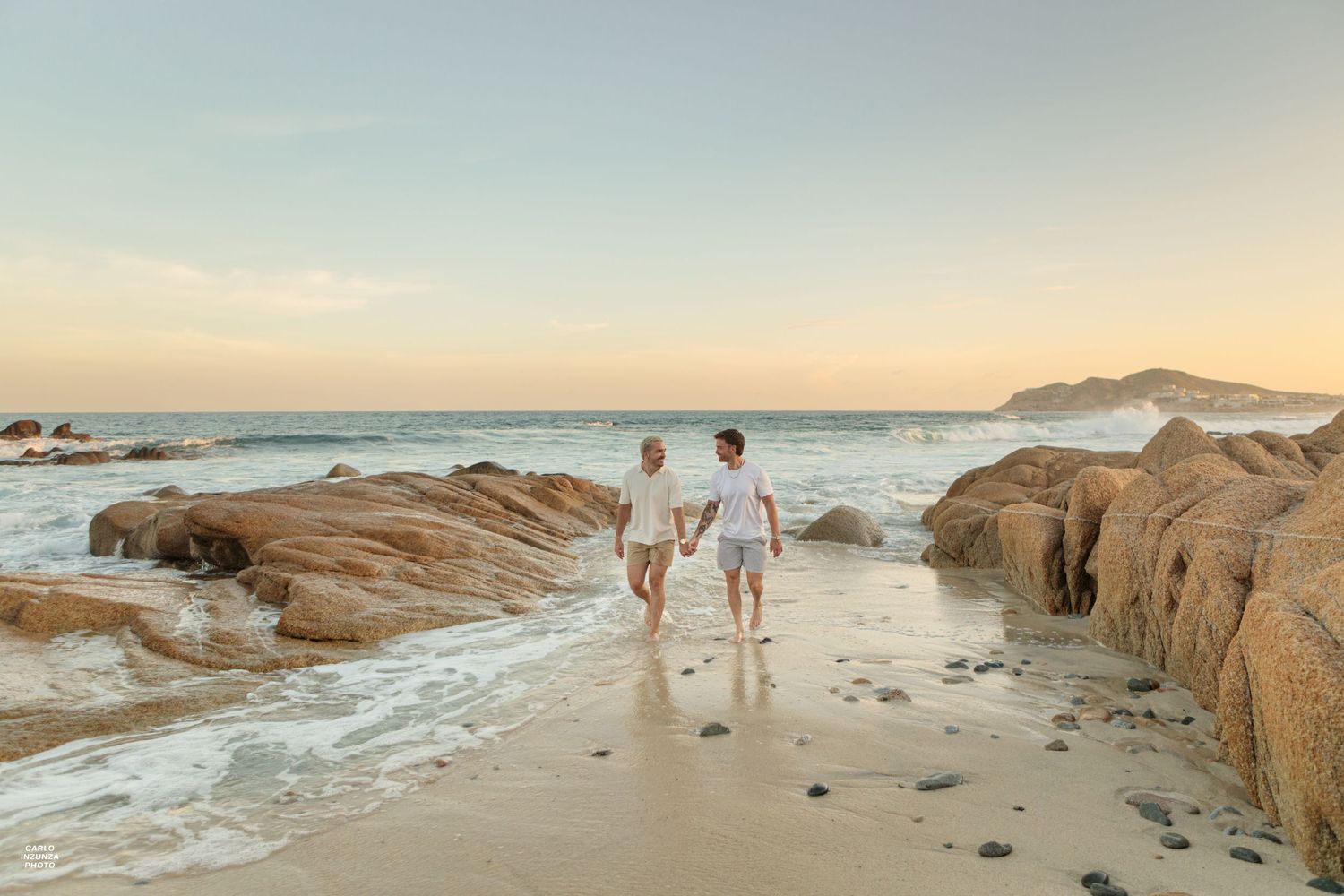 Two men walking hand in hand on sandy beach beside large rocks at golden sunset.