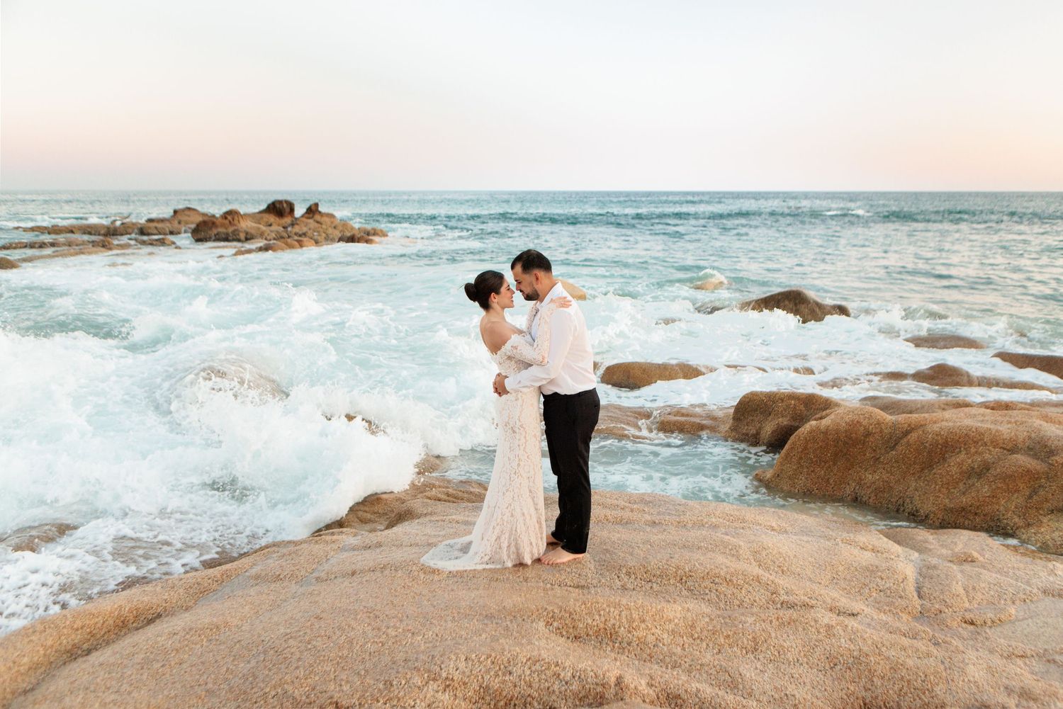 Bride and groom embrace on rocky oceanside cliffs with crashing waves at sunset during their beach wedding.