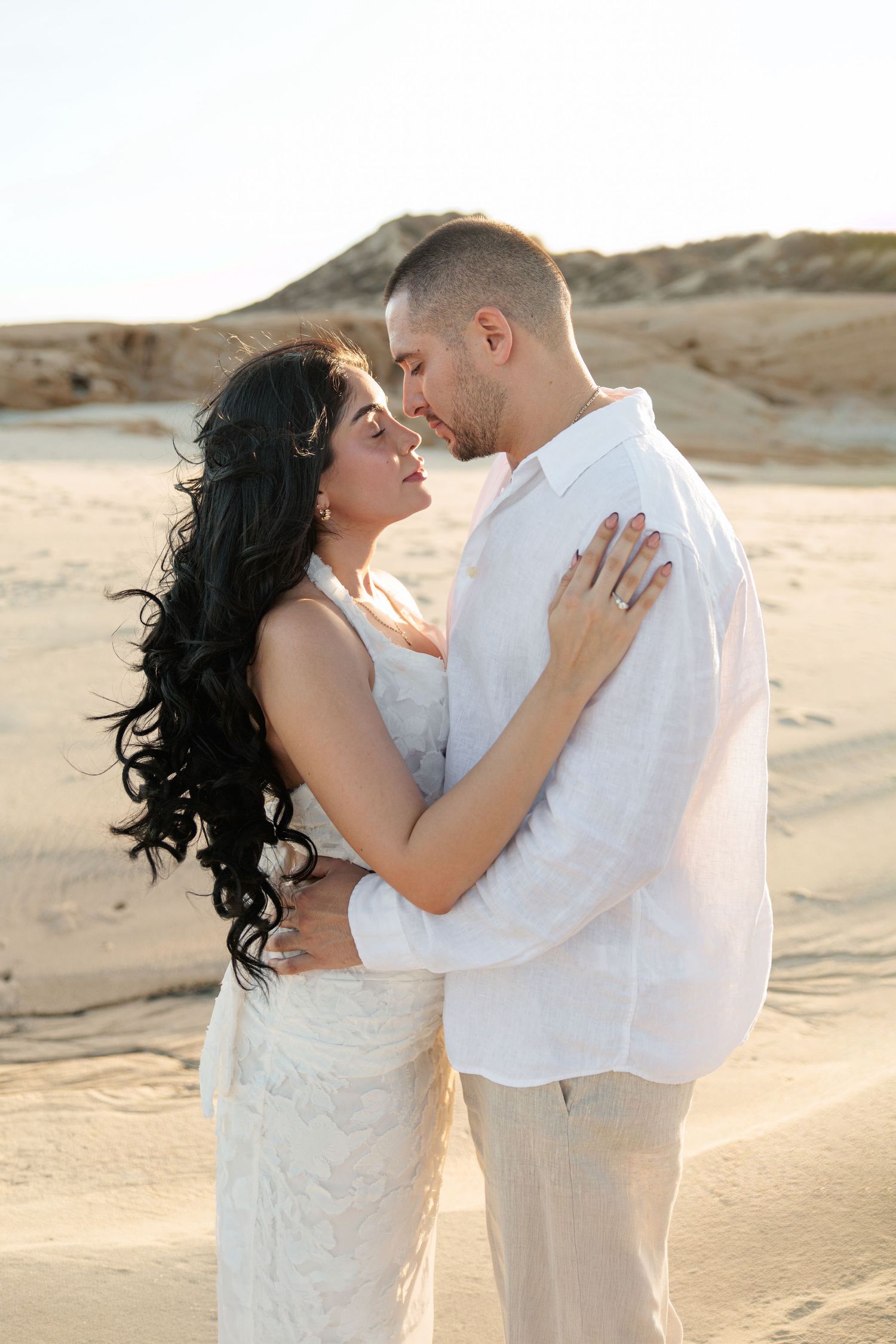 Couple in white embracing romantically on sandy desert dunes at sunset, woman with long dark hair, man in linen shirt.