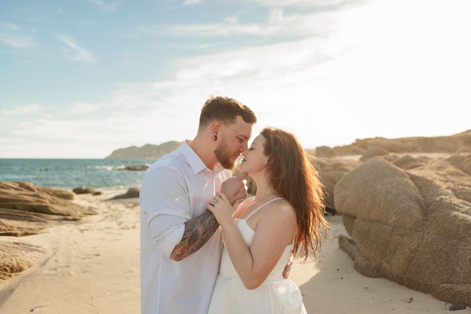 Couple sharing a kiss on a sandy beach surrounded by large rocks during a sunny golden hour session.