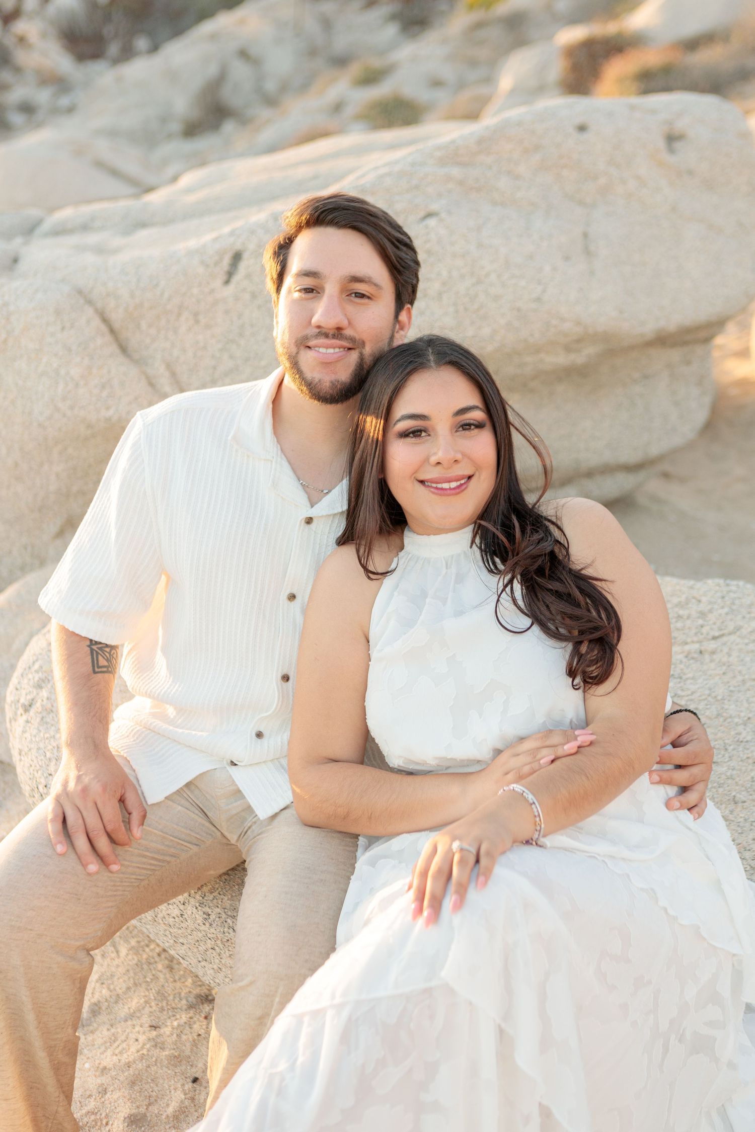 A smiling couple in white outfits sits together on rocky sand during a golden hour beach engagement photo.