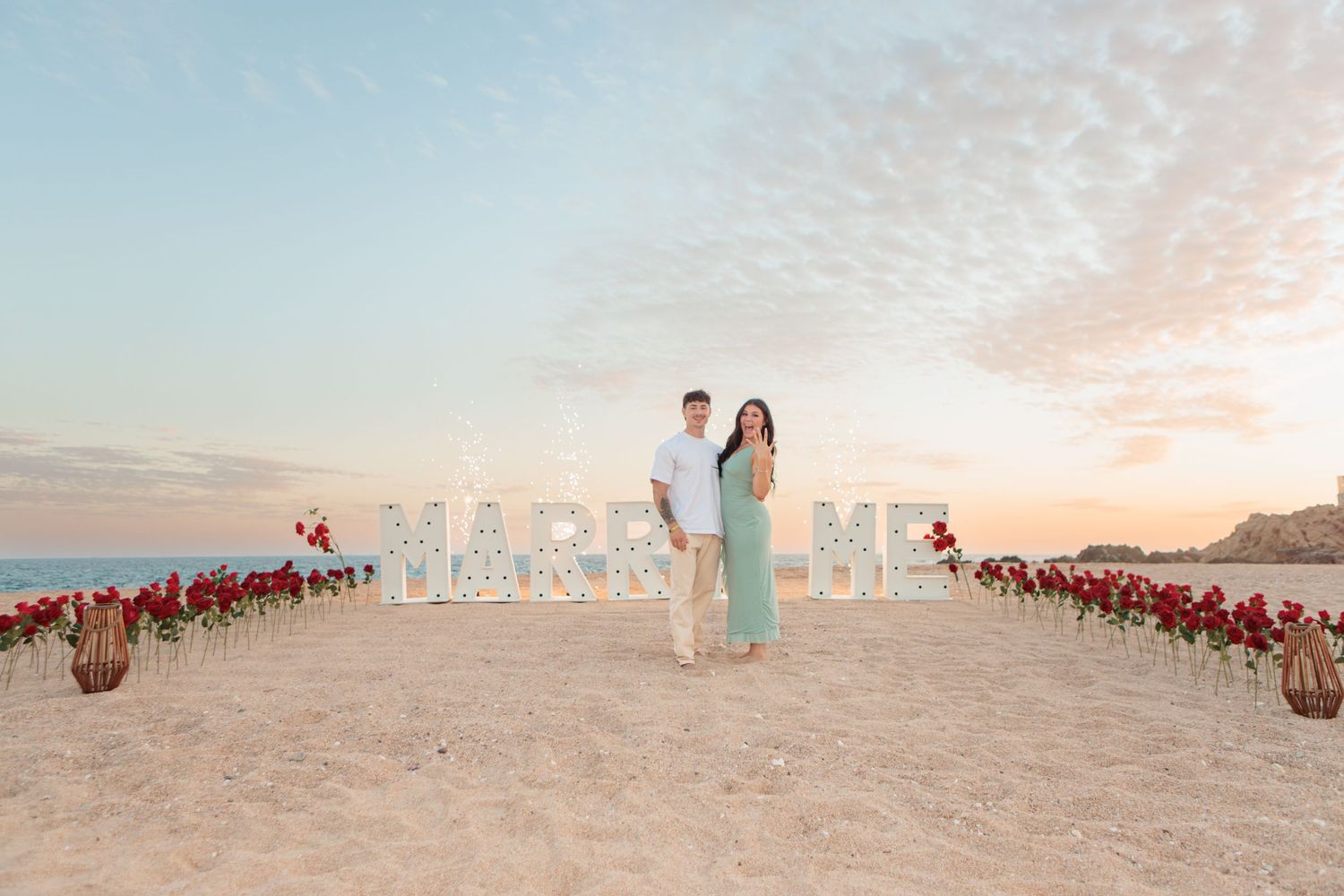 Couple posing at romantic beach proposal with lit MARRY ME sign and red roses.
