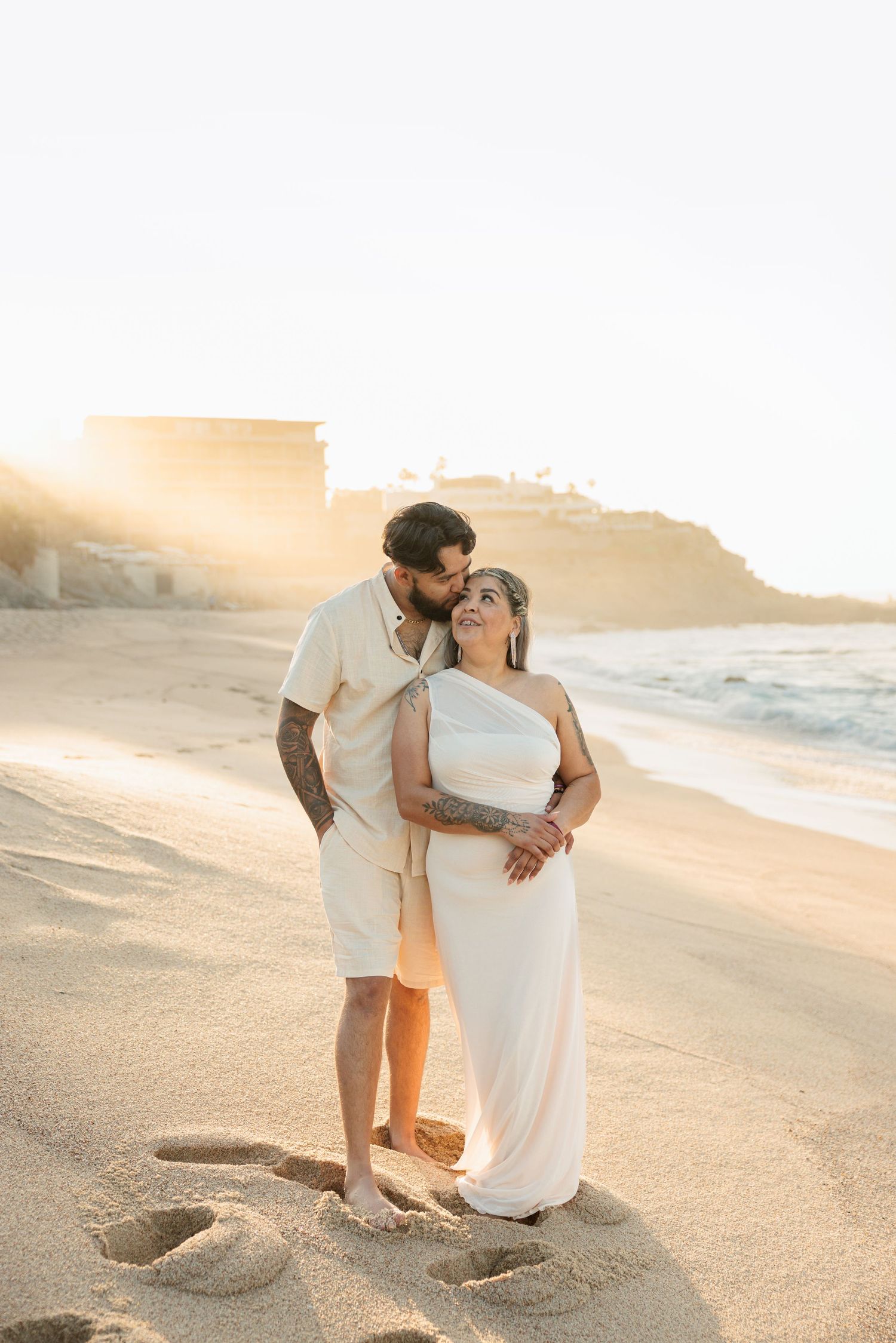 Tattooed couple sharing a tender kiss on a sunlit beach at golden hour.