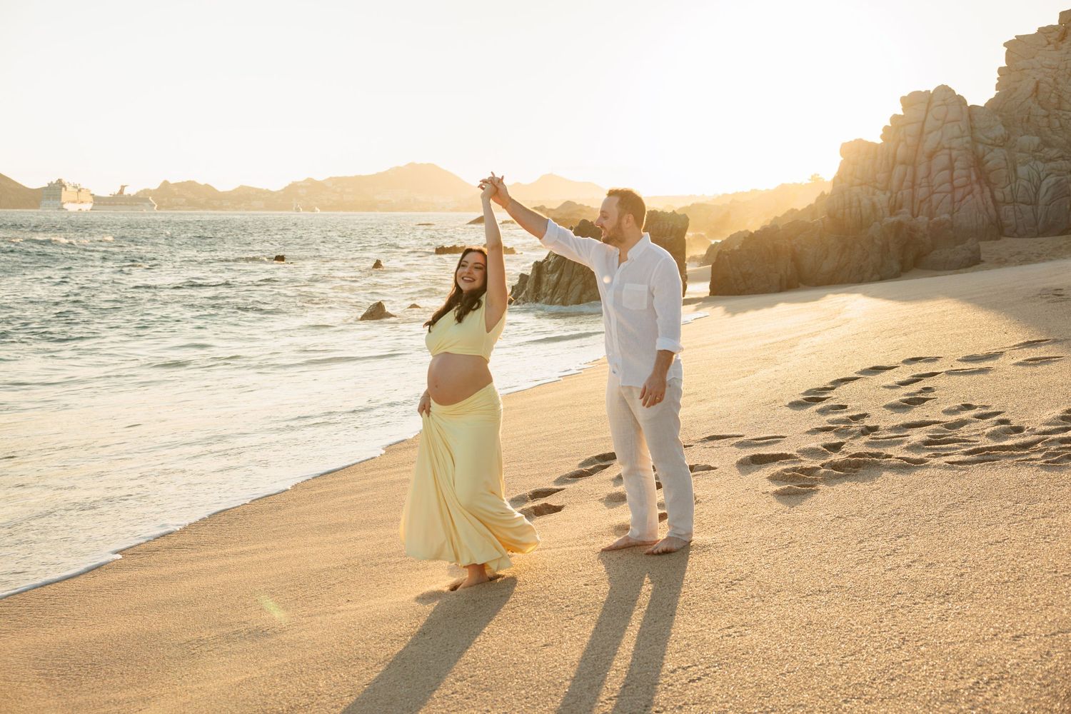 Pregnant couple dancing on sandy beach at golden sunset with rocky coastline in background.