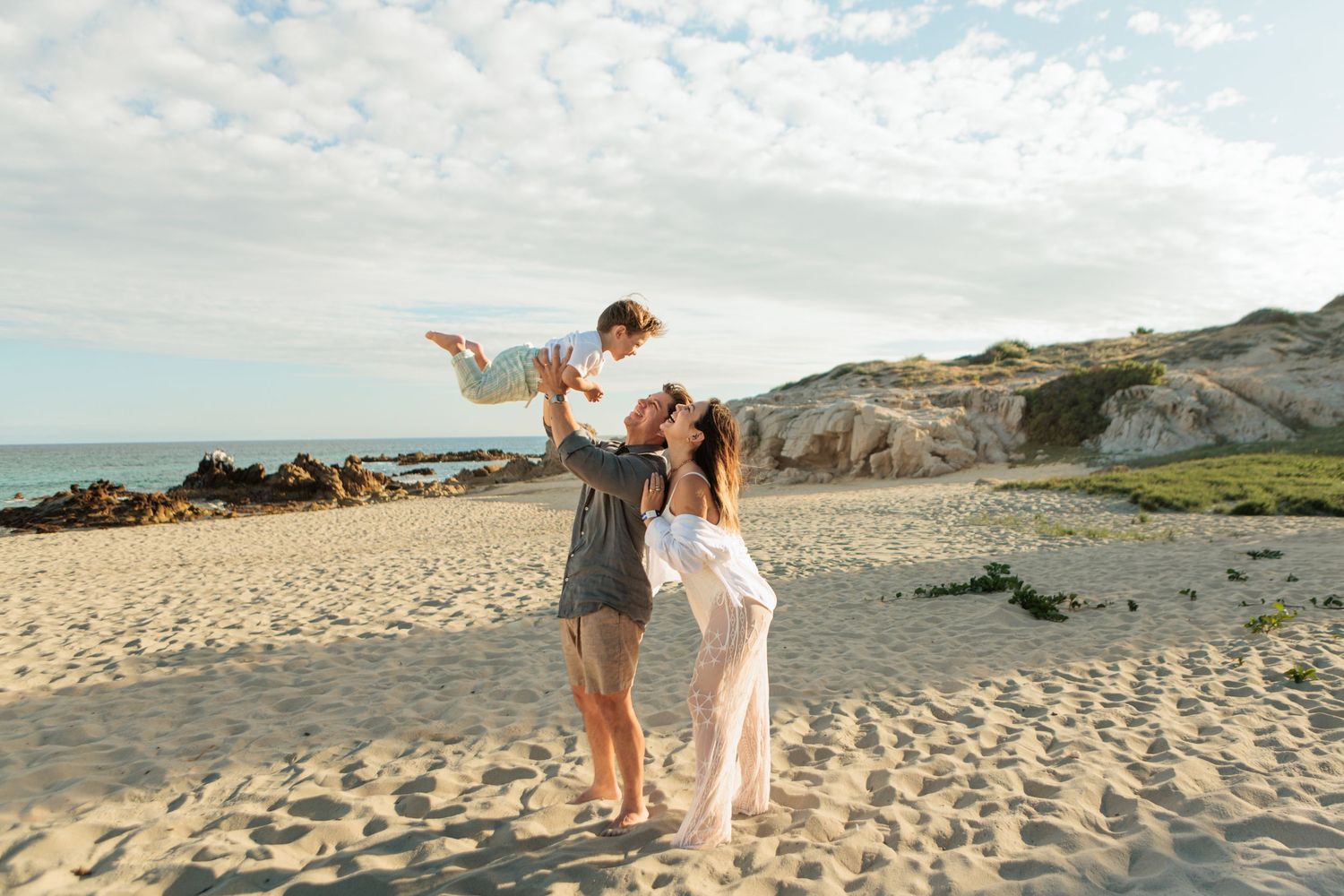 Joyful family on sandy beach at sunset, parents lifting young child against scenic coastal backdrop.