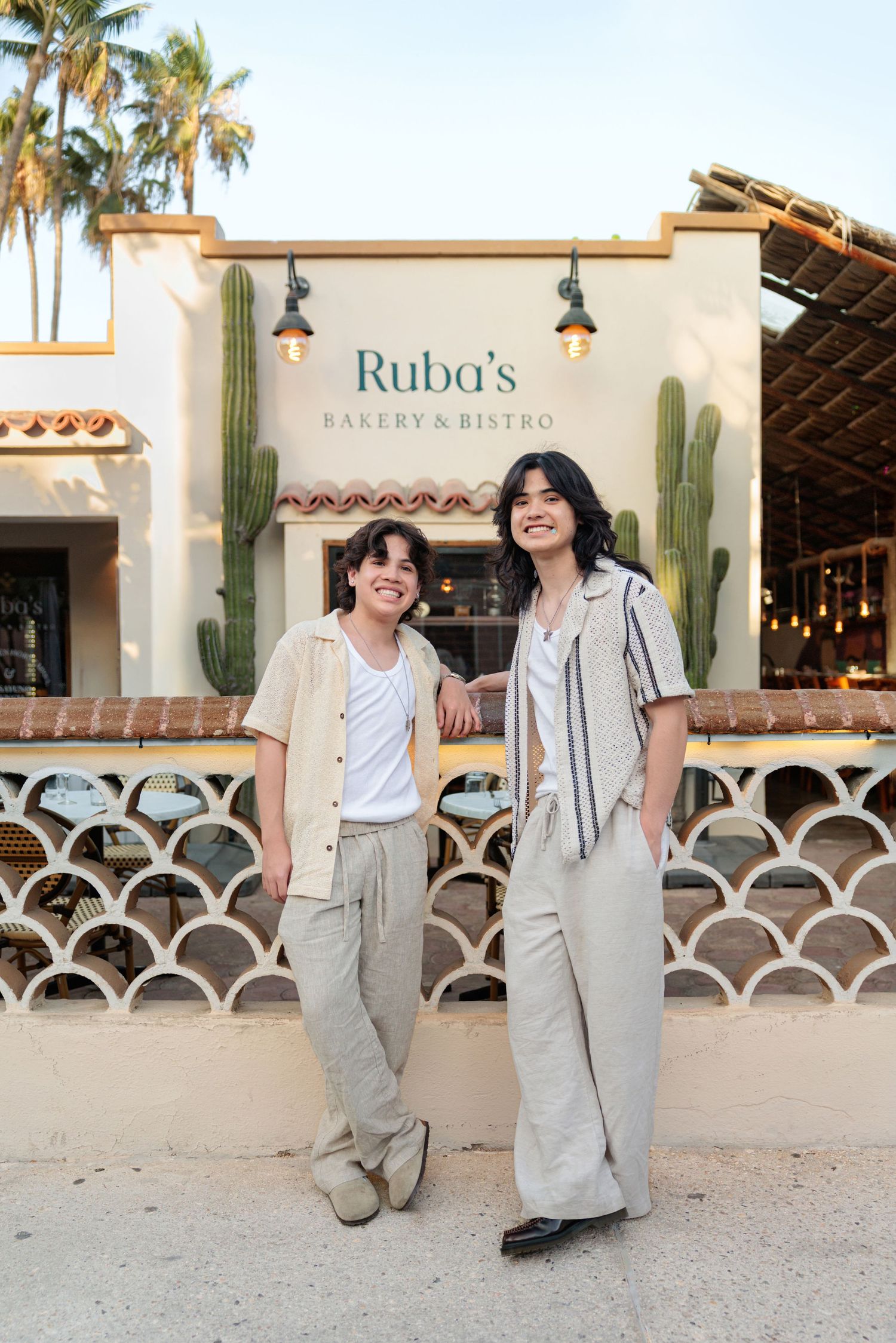 Two smiling friends pose outside Ruba's Bakery & Bistro, a charming cafe with cacti and Spanish-style architecture.