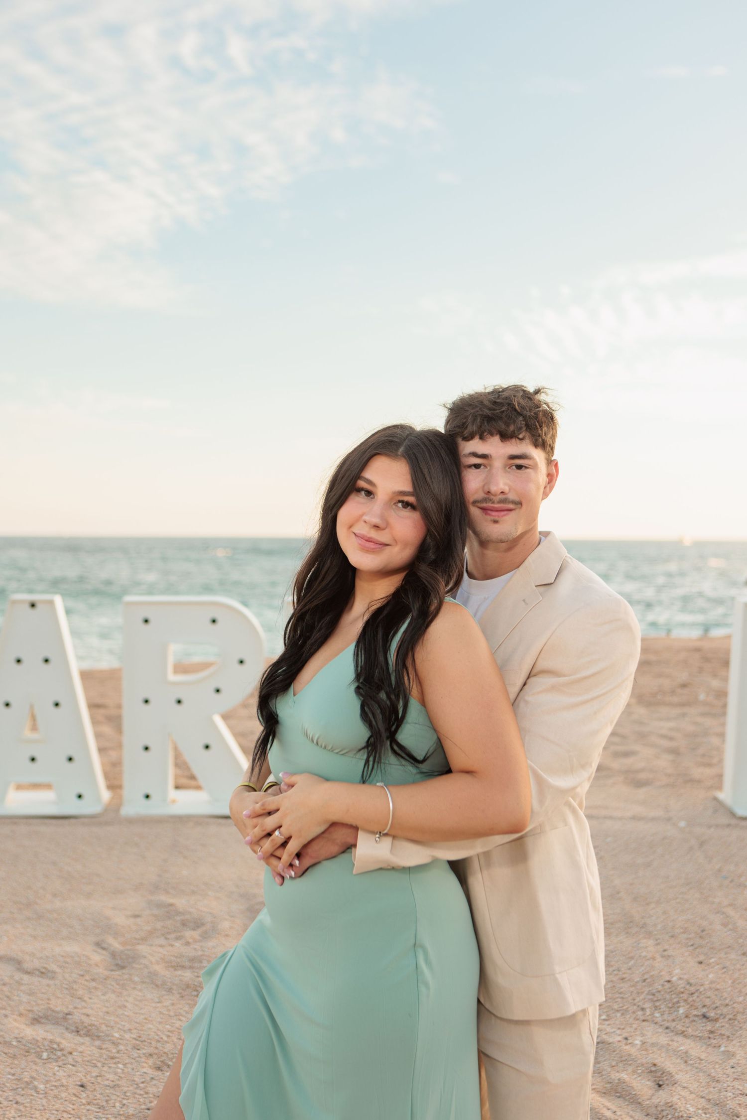 Couple embracing on a sandy beach at sunset with large marquee letters behind them.