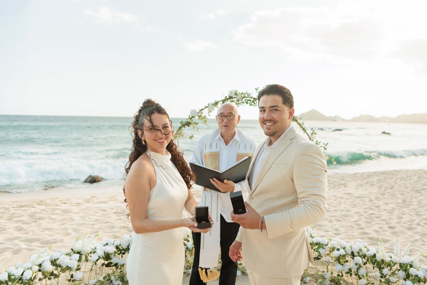 Couple exchanging wedding vows on a beach with an officiant and floral arch backdrop at a destination wedding.