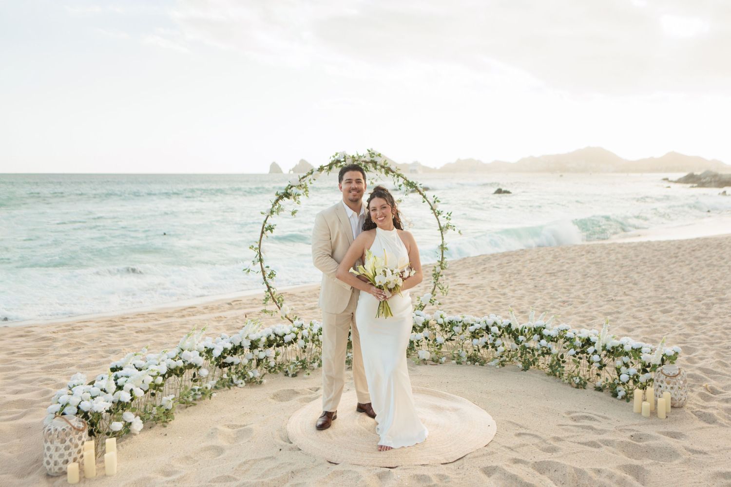Bride and groom standing under circular floral arch on beach, white flowers, ocean waves in background at sunset.