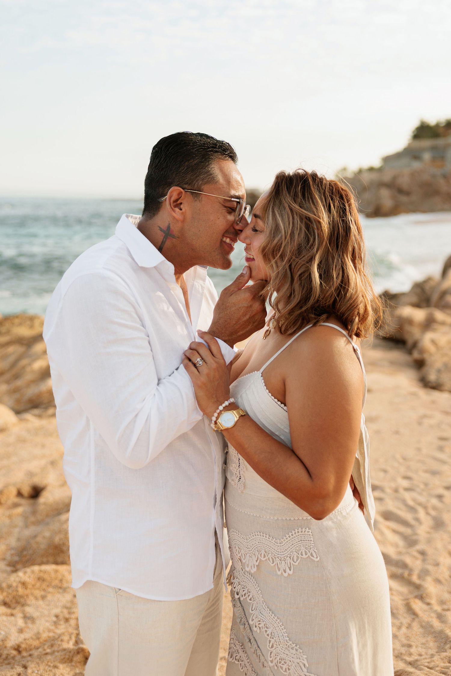 Couple in white outfits sharing a romantic moment on a rocky beach at sunset.