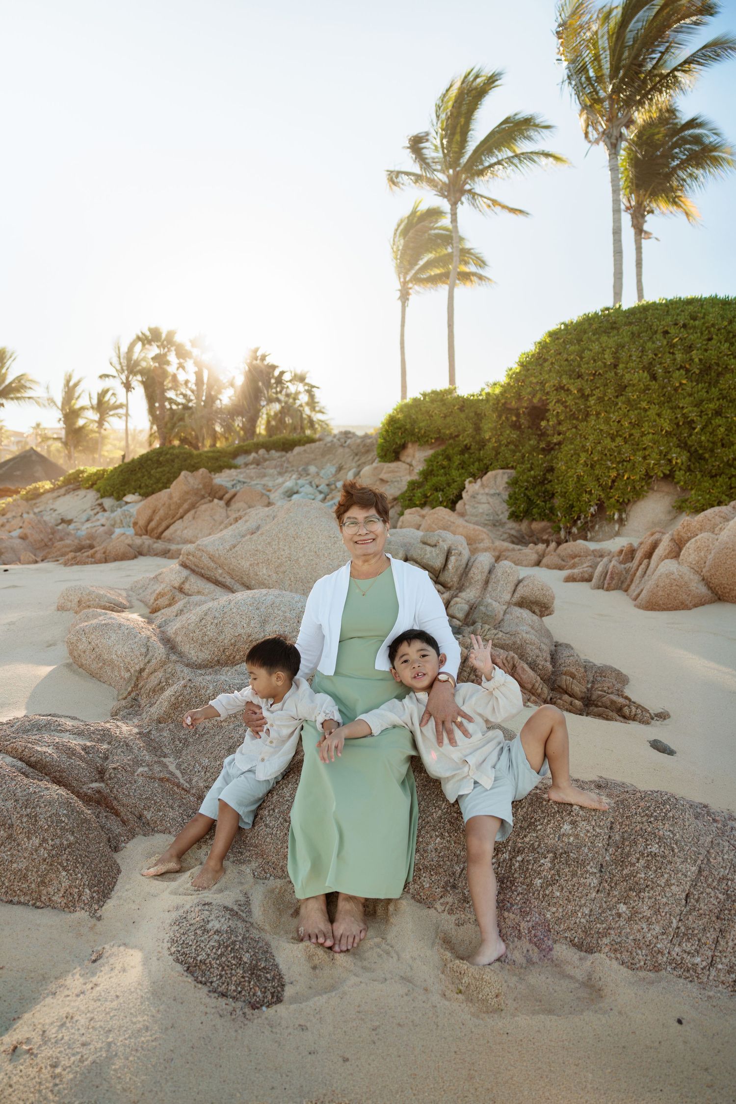 Woman with two children sitting on rocks at a sandy beach with palm trees and golden sunset light.