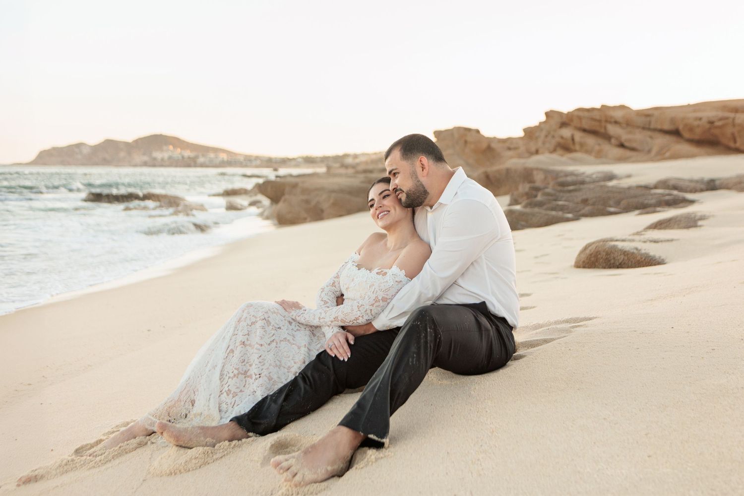 A couple in wedding attire sits romantically on sandy beach at sunset, with ocean waves and rocky cliffs in background.