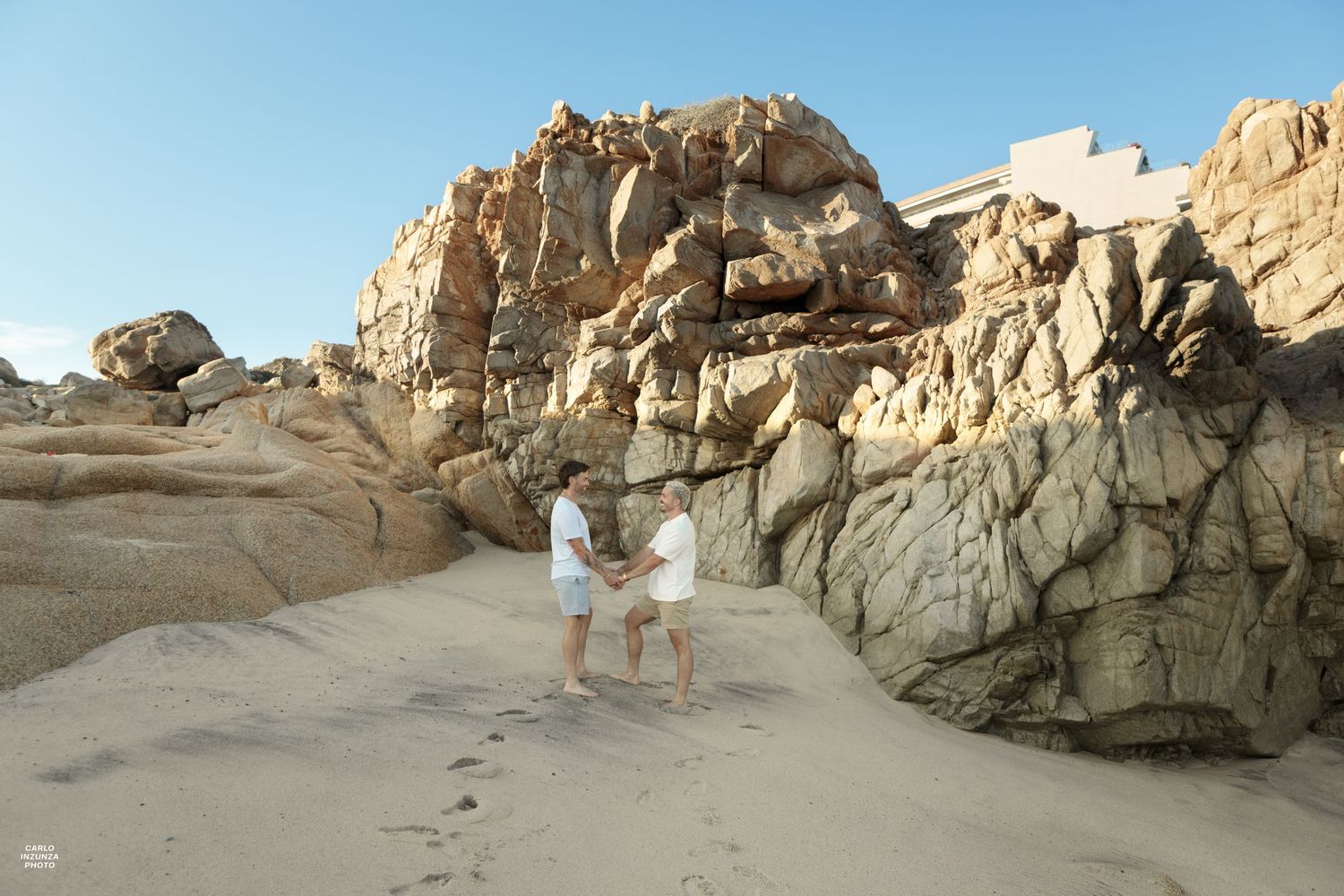 Two men holding hands on sandy beach path between large rocky boulders in bright sunlight.