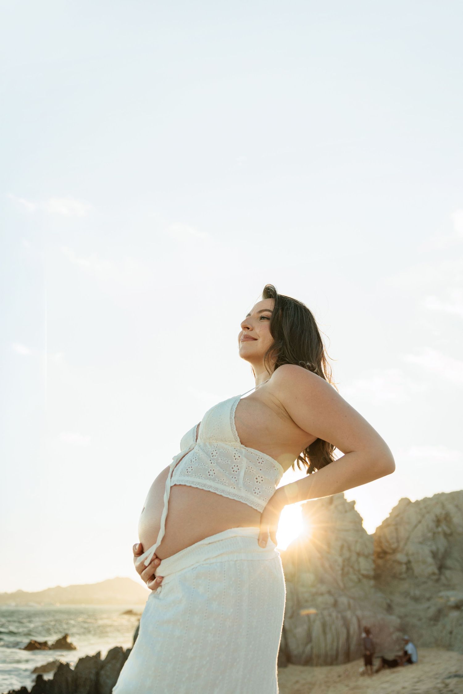 Pregnant woman in white lace top and skirt poses on rocky beach during golden sunset.