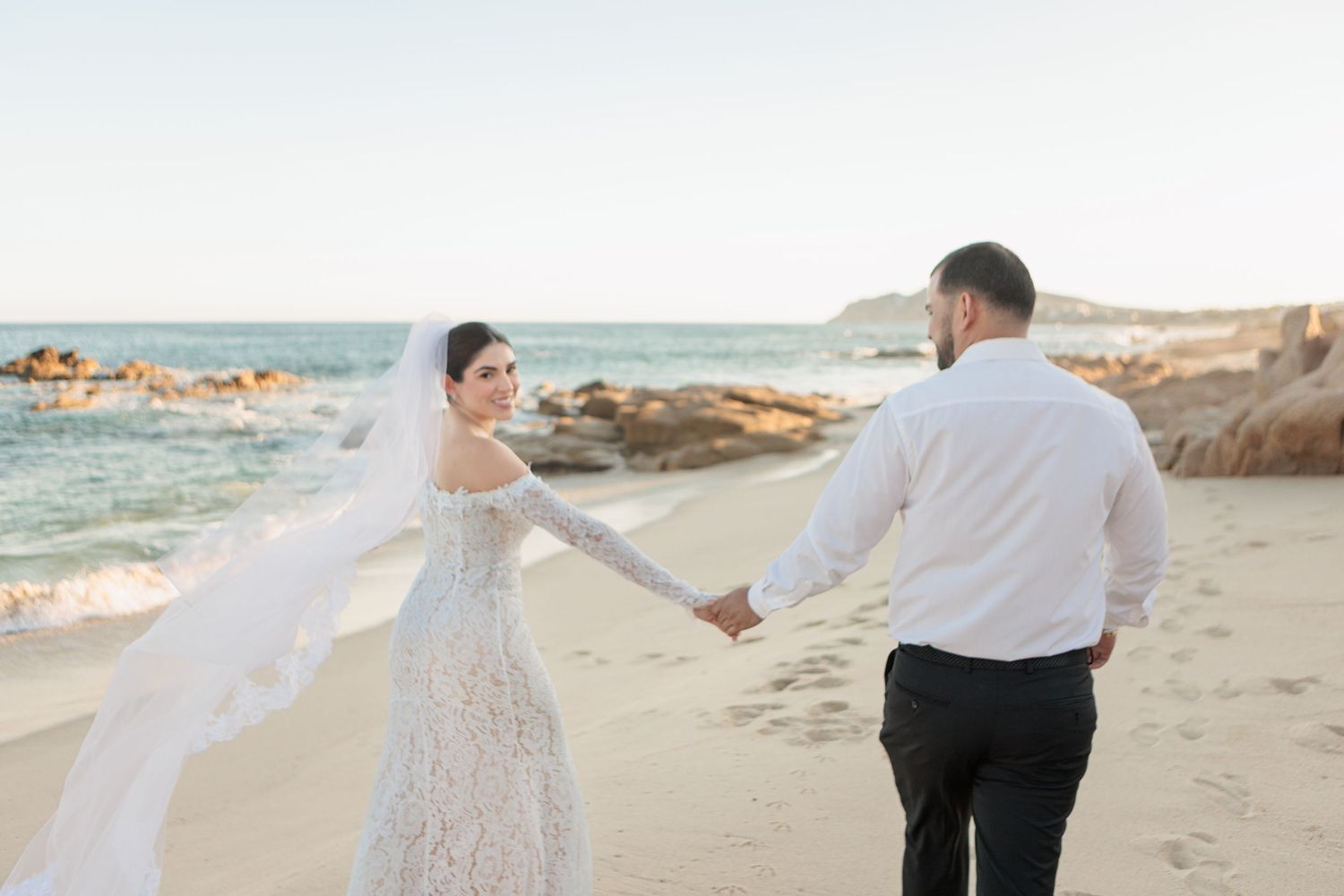 Bride in lace gown with flowing veil holds groom's hand walking on sandy beach with rocky shoreline at sunset.