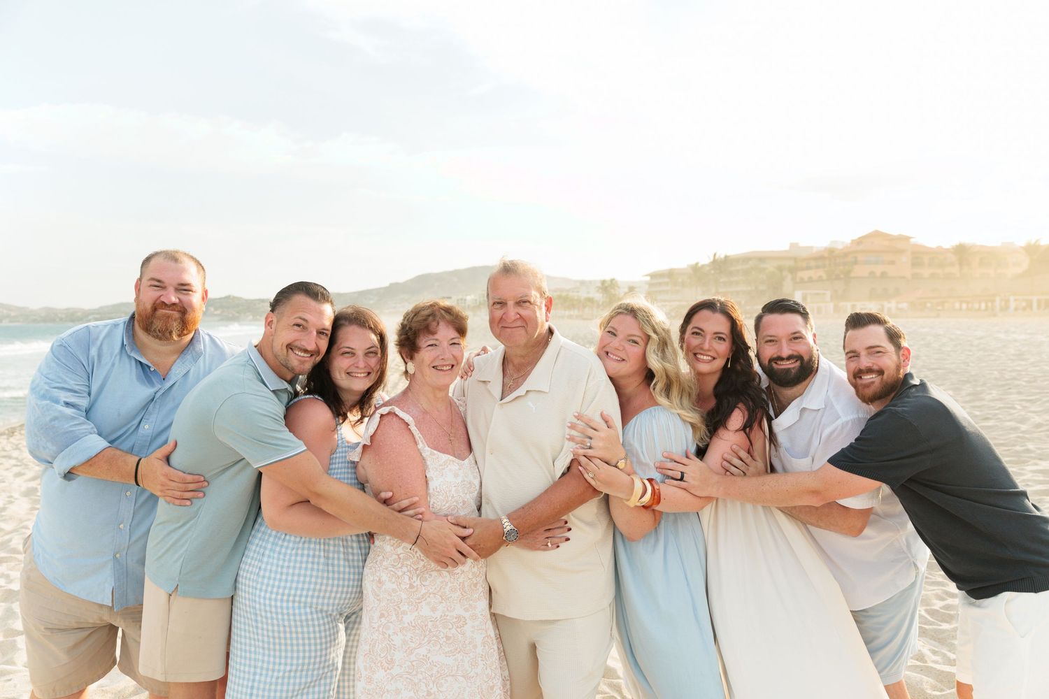 Large family group hugs together on sandy beach during sunny outdoor wedding celebration photo.