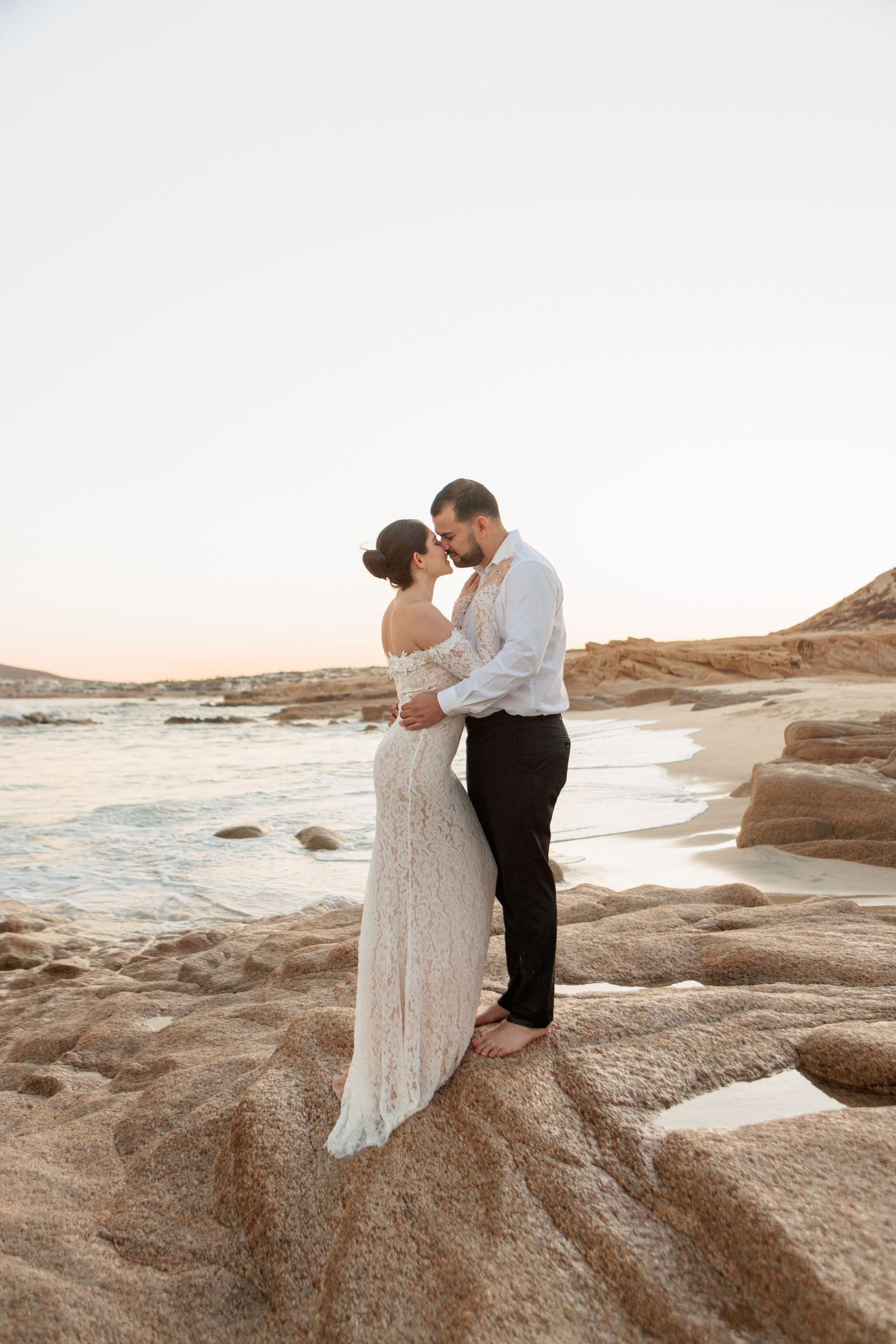 Bride and groom share a romantic moment on rocky coastal shore at sunset, bride wearing elegant lace off-shoulder gown.