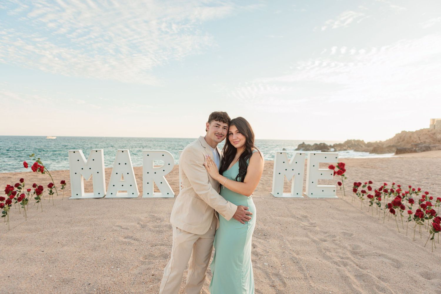 Couple hugging on beach in front of illuminated MARRY ME marquee letters with red roses.