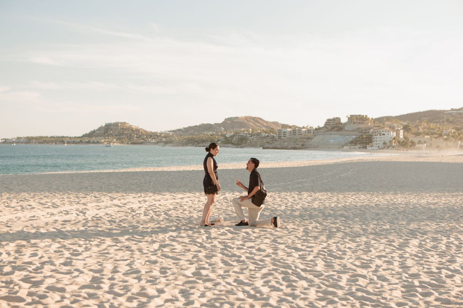 Man proposing on one knee to his partner on a white sandy beach.