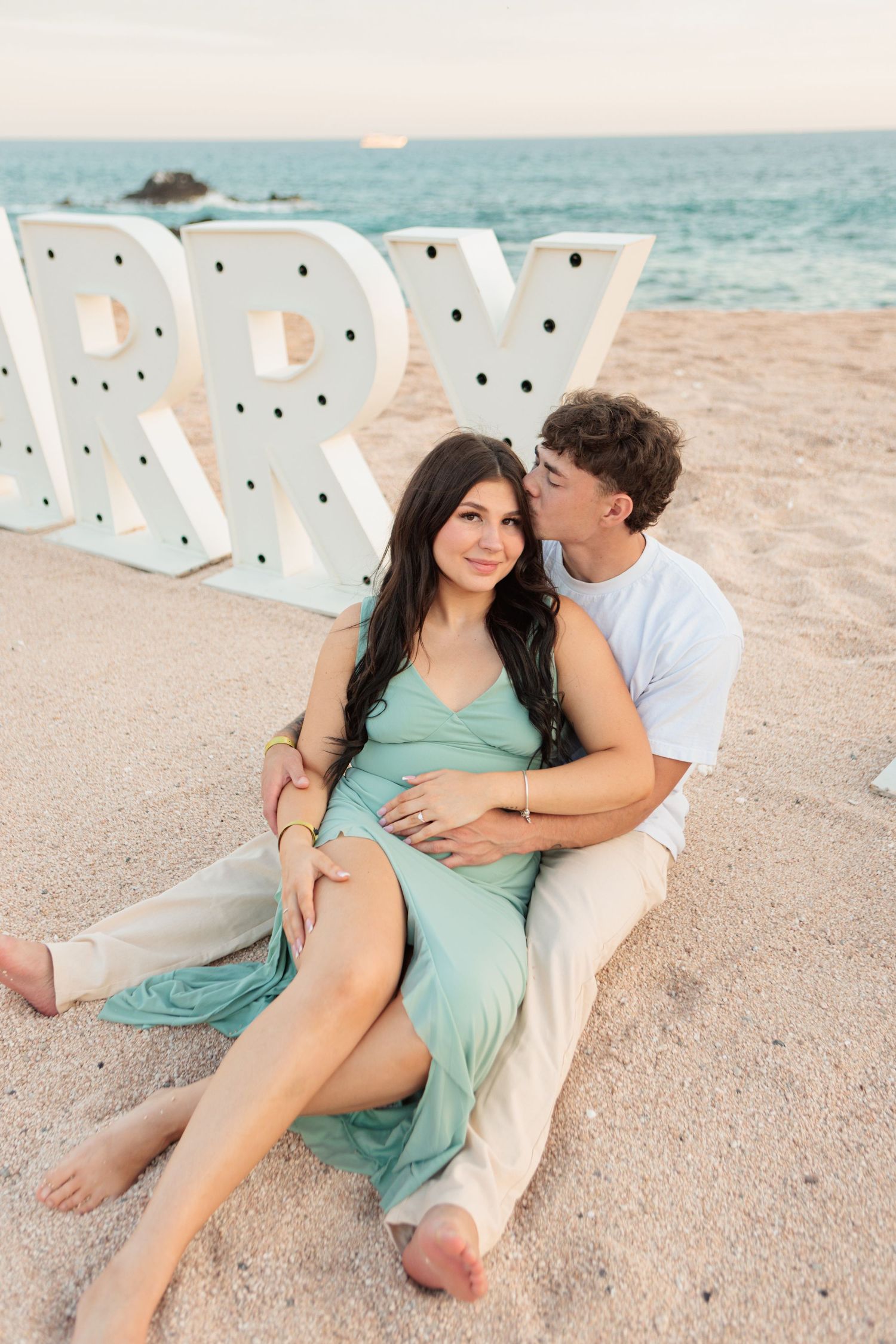 Couple sitting on sand in front of MARRY ME marquee letters at beach sunset.