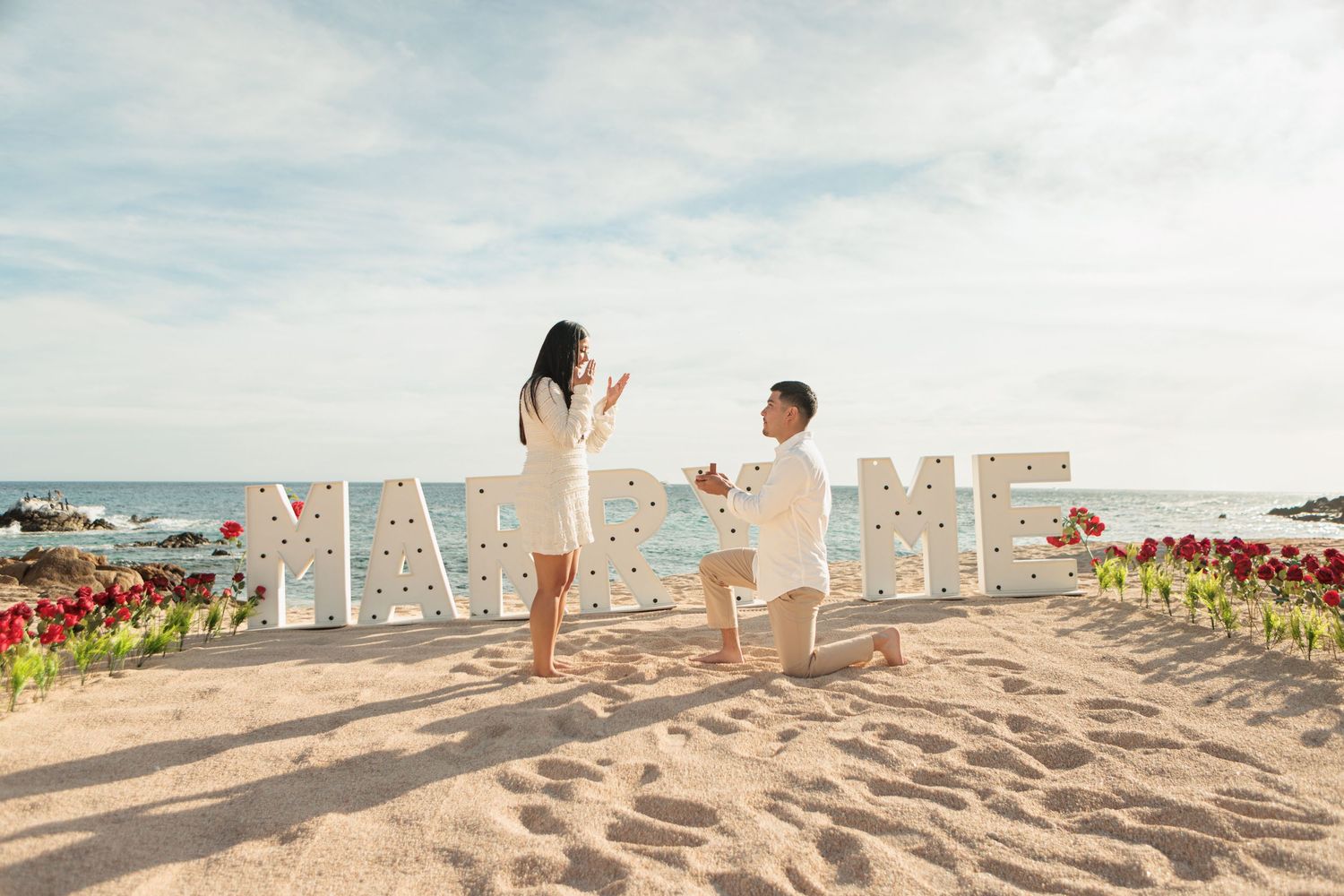 Man proposing to woman on beach with illuminated MARRY ME sign and red roses at sunset.
