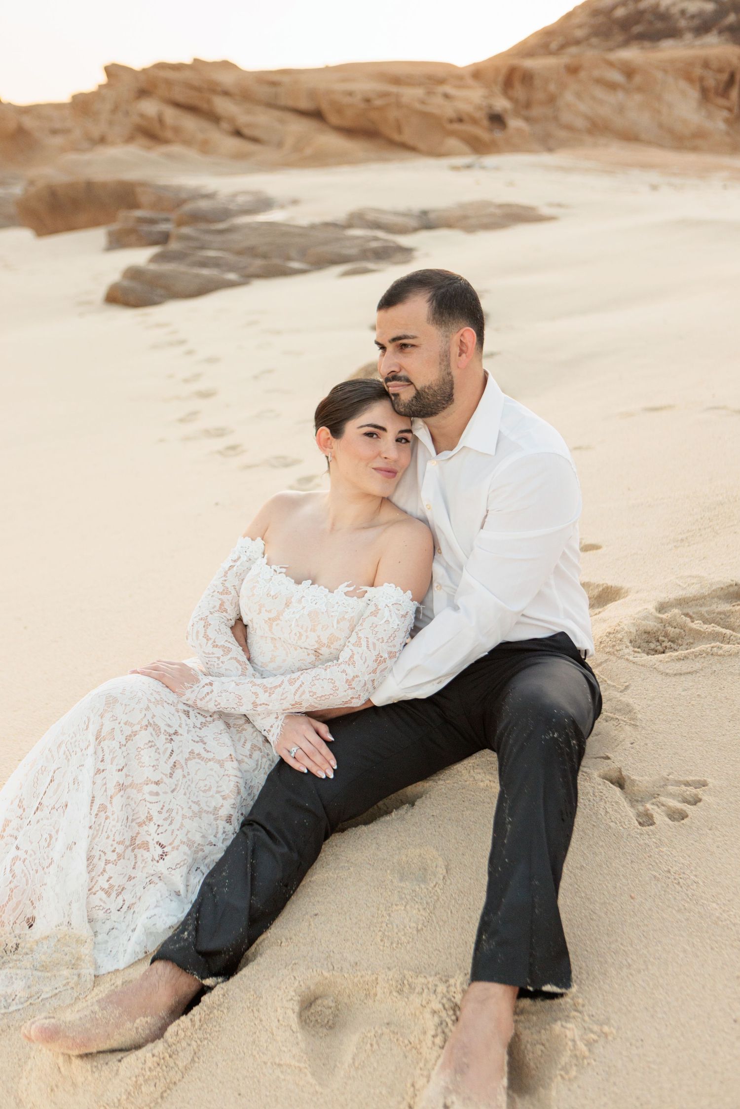 Bride and groom sit together on sandy dunes at sunset, bride in lace gown, groom in white shirt.