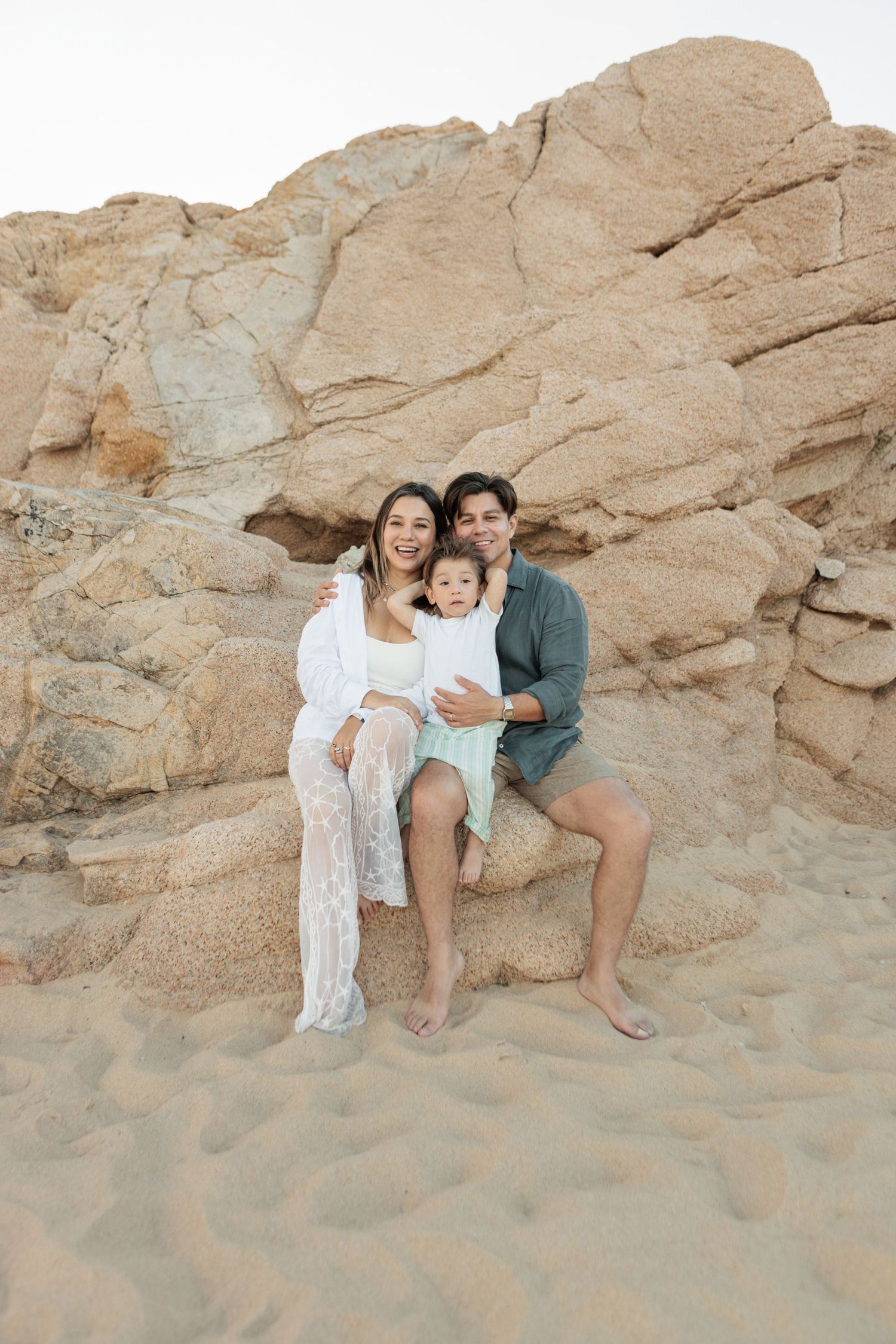 Happy family of three sitting together on sandy rocks at the beach, smiling for a portrait.