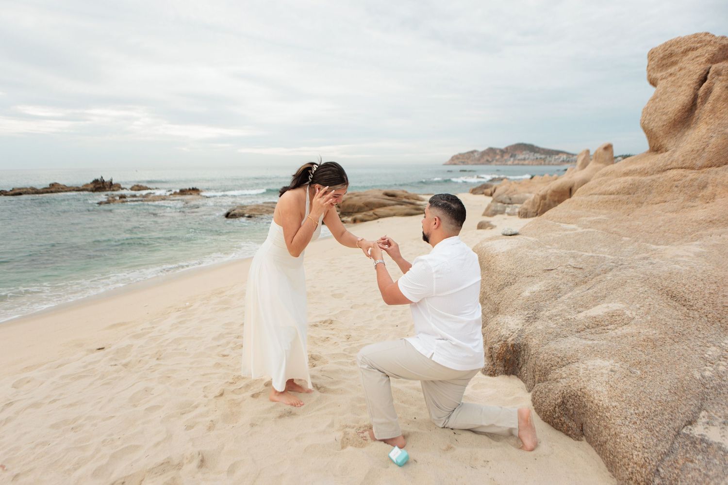 Man proposing on one knee to woman in white dress on a rocky beach.