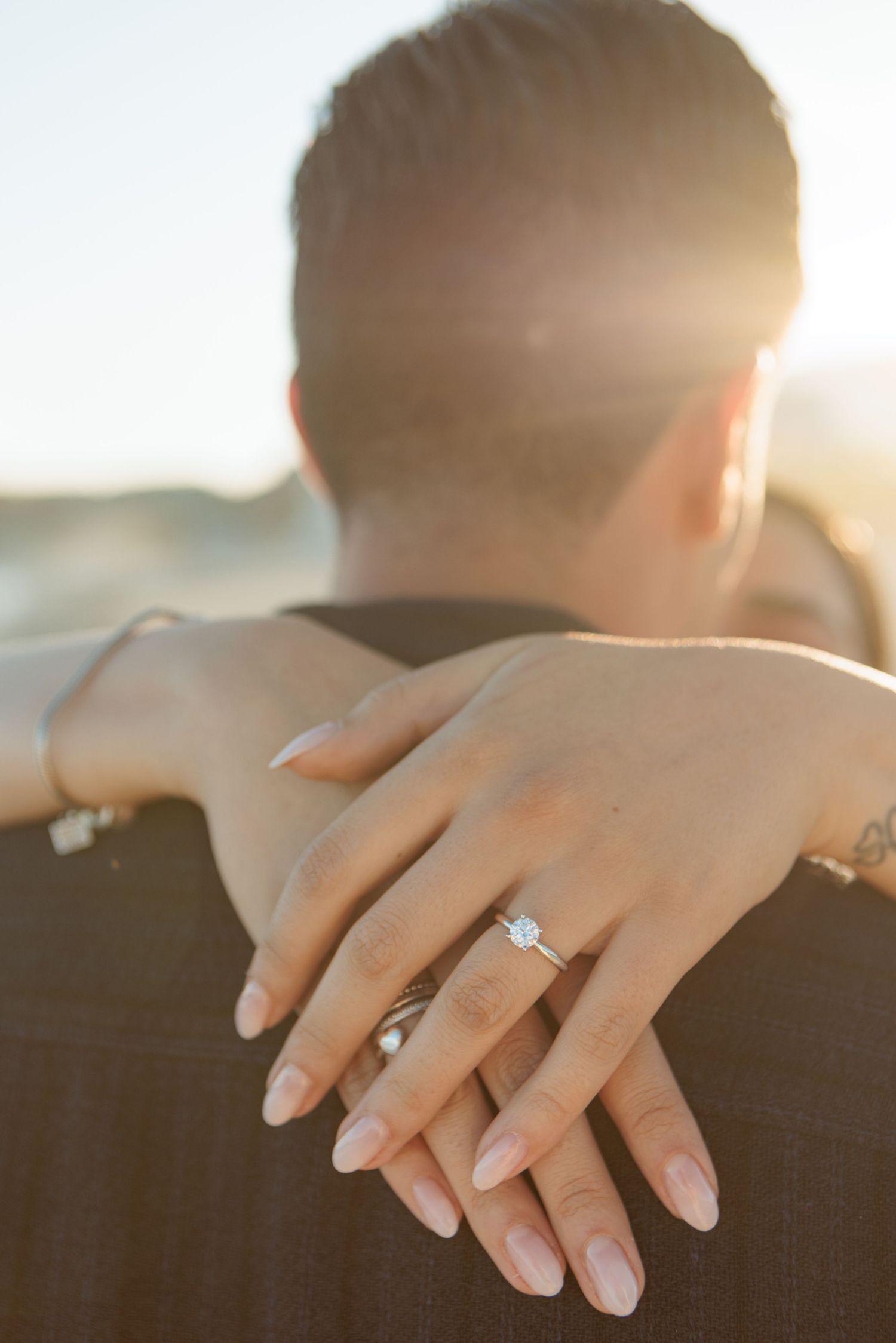 Close-up of a diamond engagement ring on a woman's hand resting on a man's shoulder at sunset.