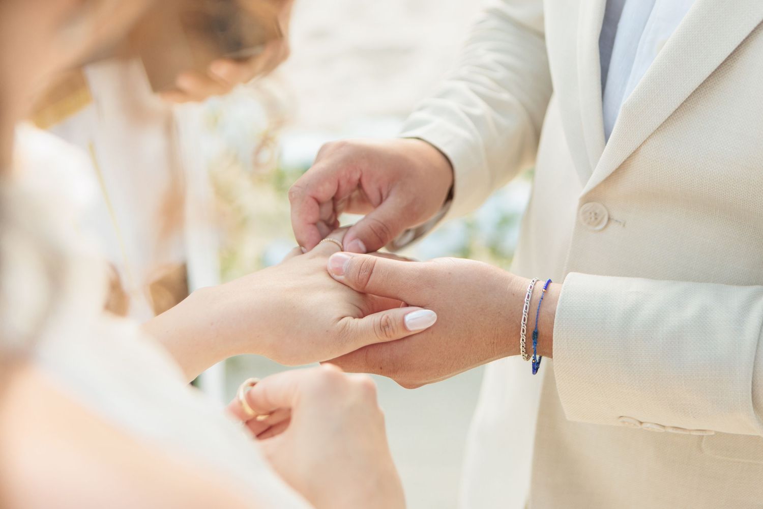 Groom in white suit places wedding ring on bride's finger during an outdoor ceremony in soft natural light.