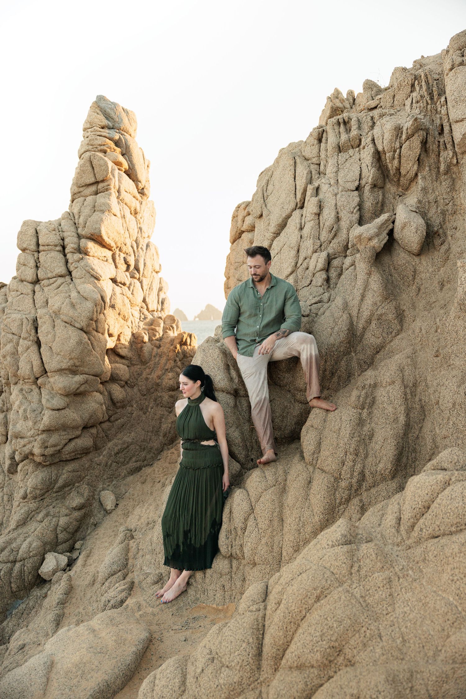 Couple posing on rocky coastal cliffs, woman in green dress standing below man seated on rocks above her.