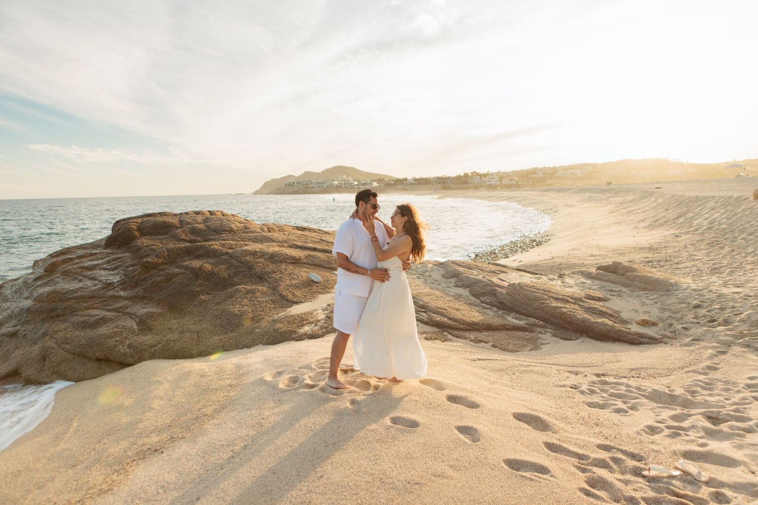 Couple in white outfits embracing on sandy beach at golden sunset with ocean and rocks backdrop.