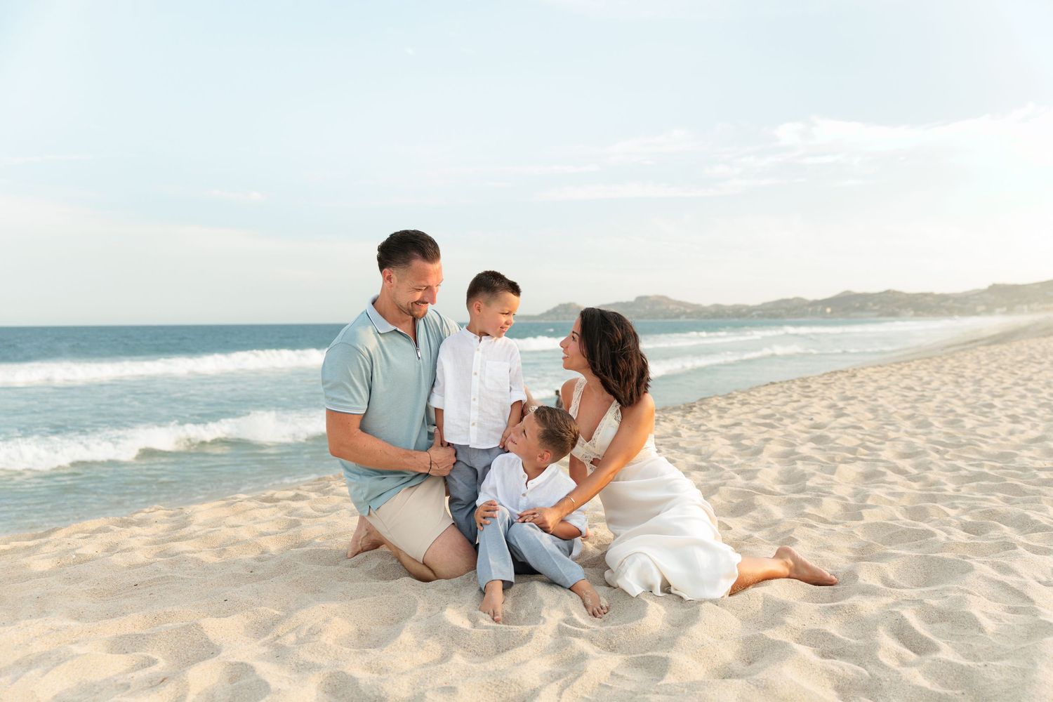 Family of four sitting together on a sandy beach, smiling and enjoying a sunny day by the ocean waves.
