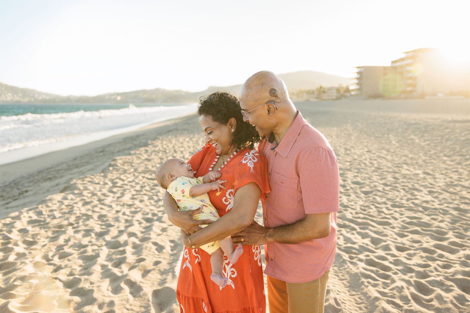 Happy couple holding newborn baby on sunny beach with ocean waves and resort in background.