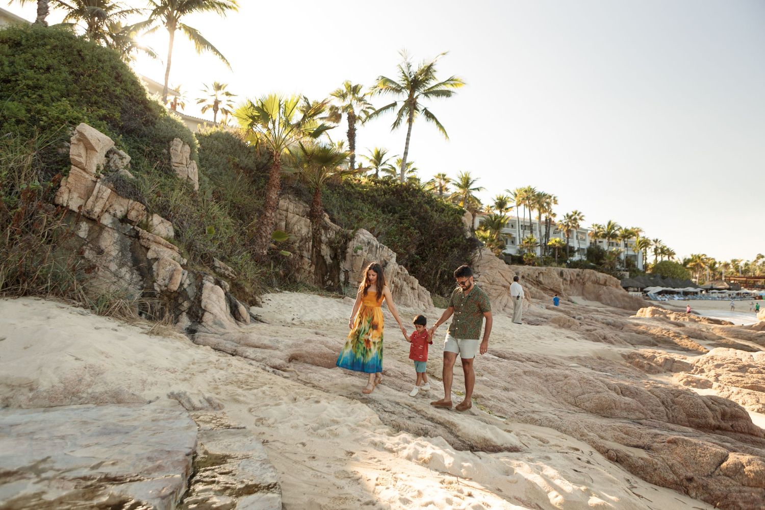 Family of three walking along rocky coastal path lined with tropical palm trees.