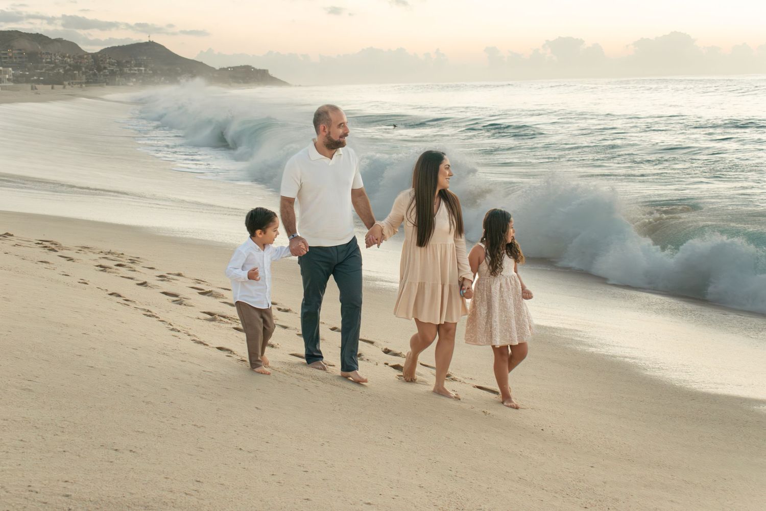Family of four walking hand-in-hand on sandy beach with crashing waves at sunset.