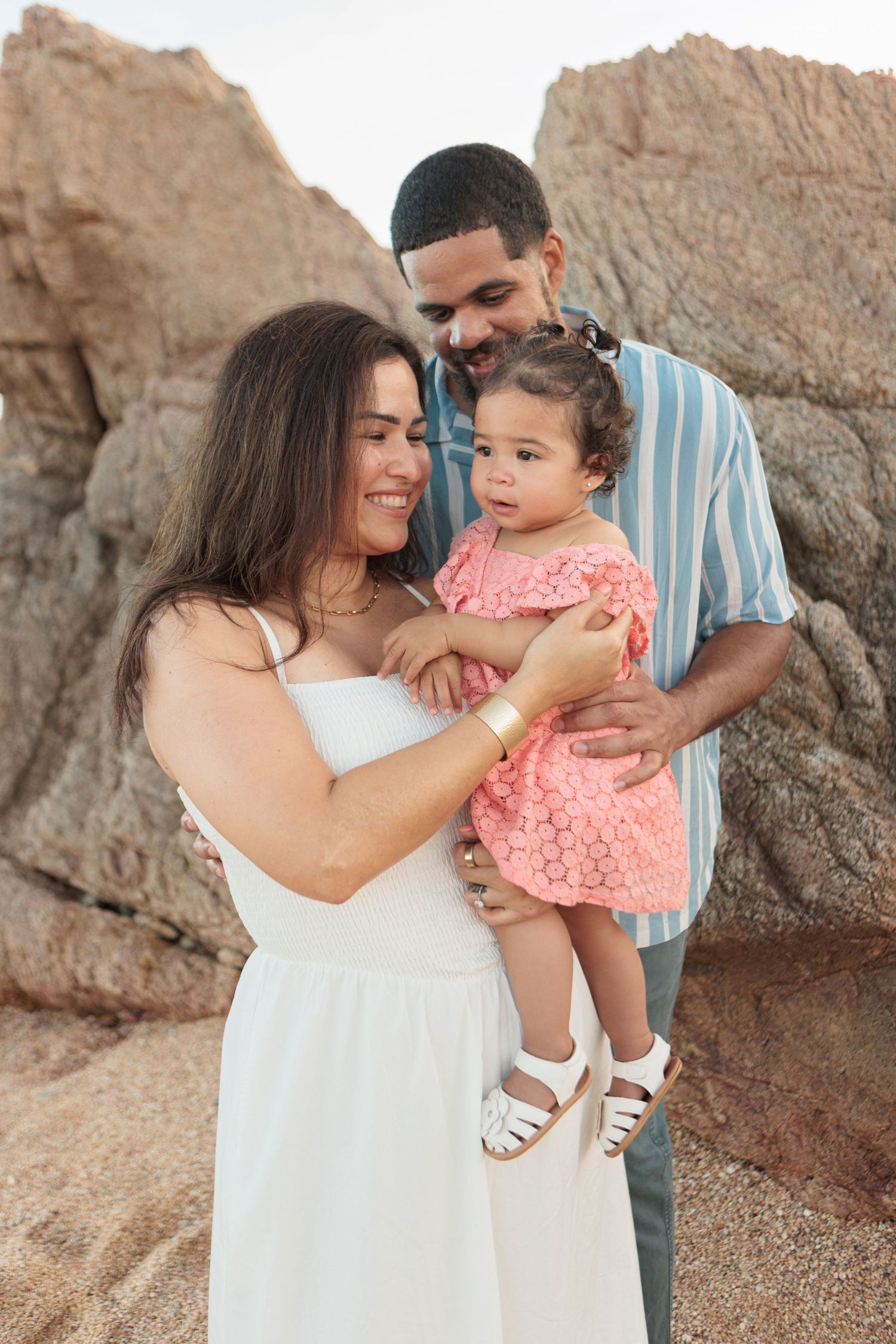 Happy family with mother in white dress and father holding baby girl in pink on a rocky beach.