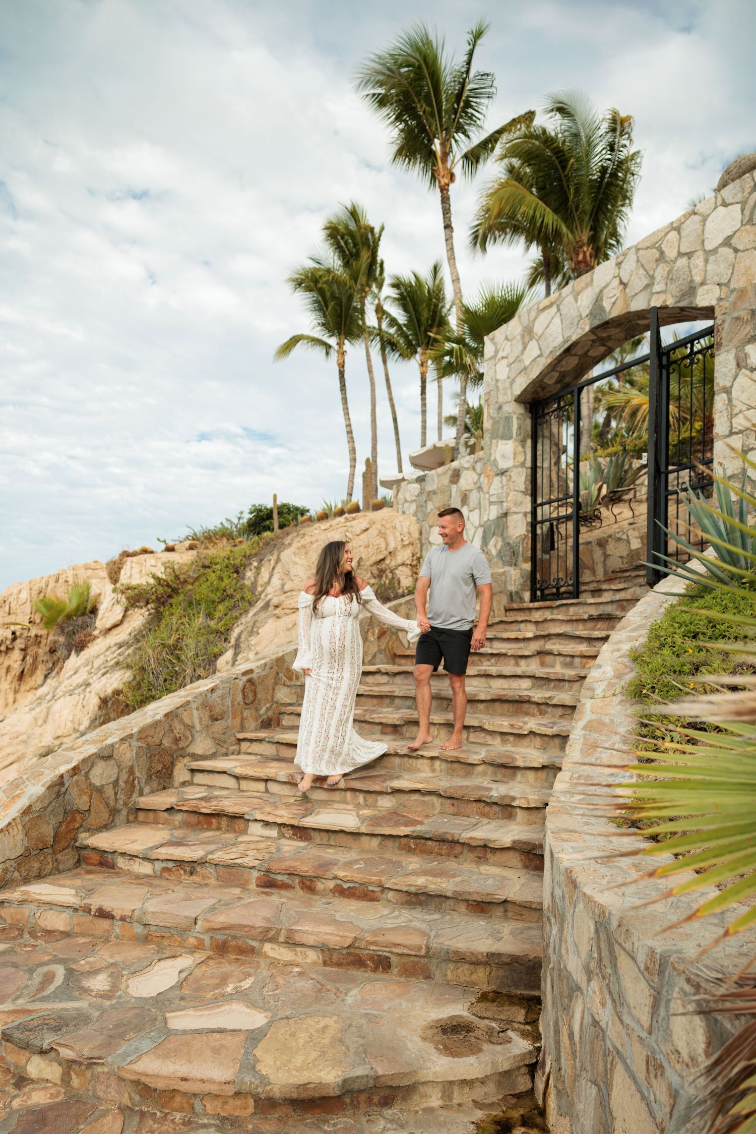 Couple holding hands on stone steps with palm trees and a stone archway at a tropical resort.
