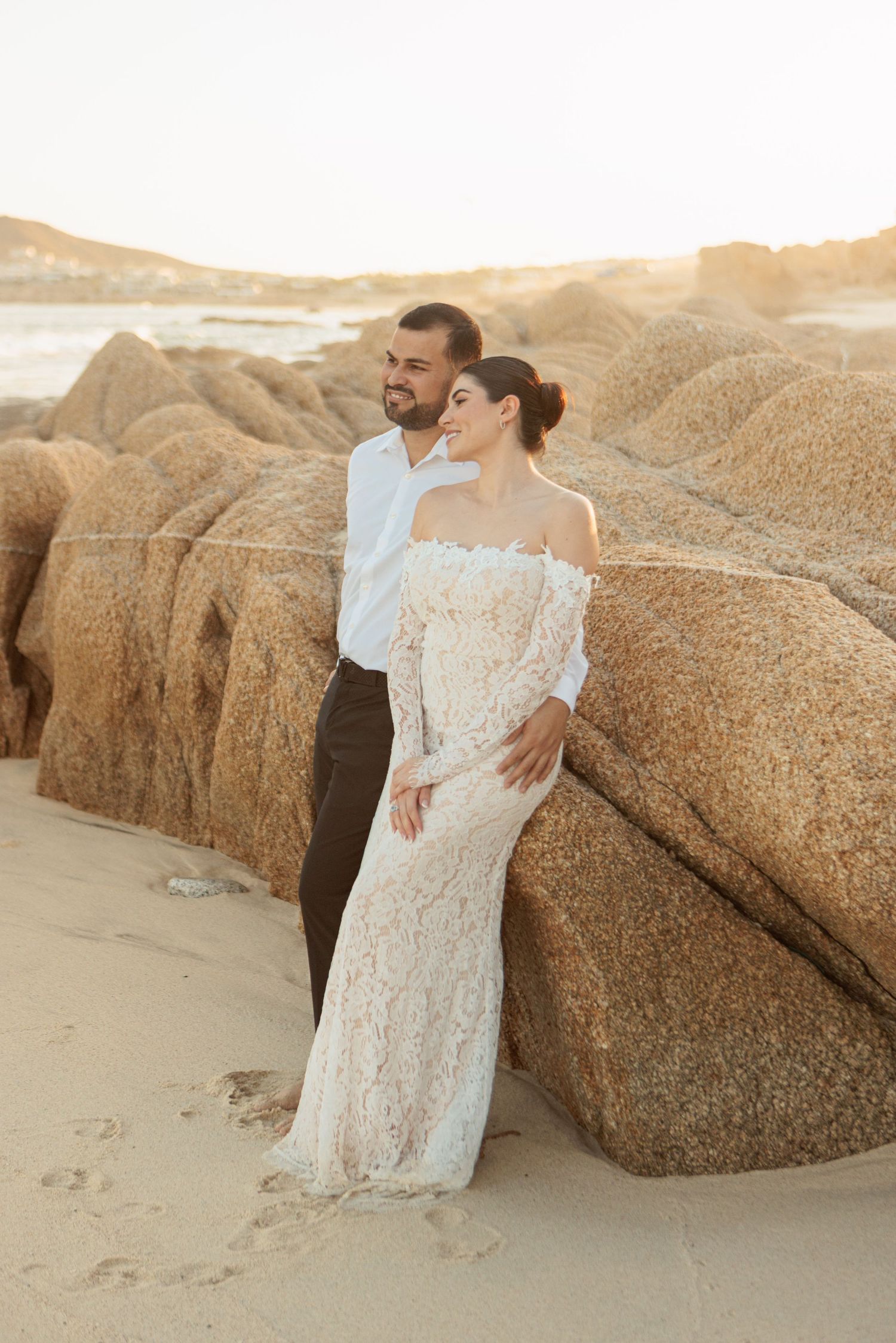 Bride in off-shoulder lace gown leans against large rocks as groom embraces her during golden hour beach shoot.