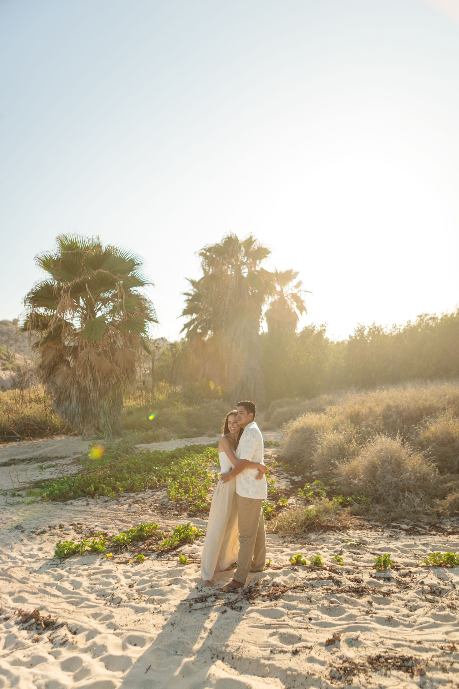 Couple embraces on sandy beach surrounded by palm trees in golden sunlight.