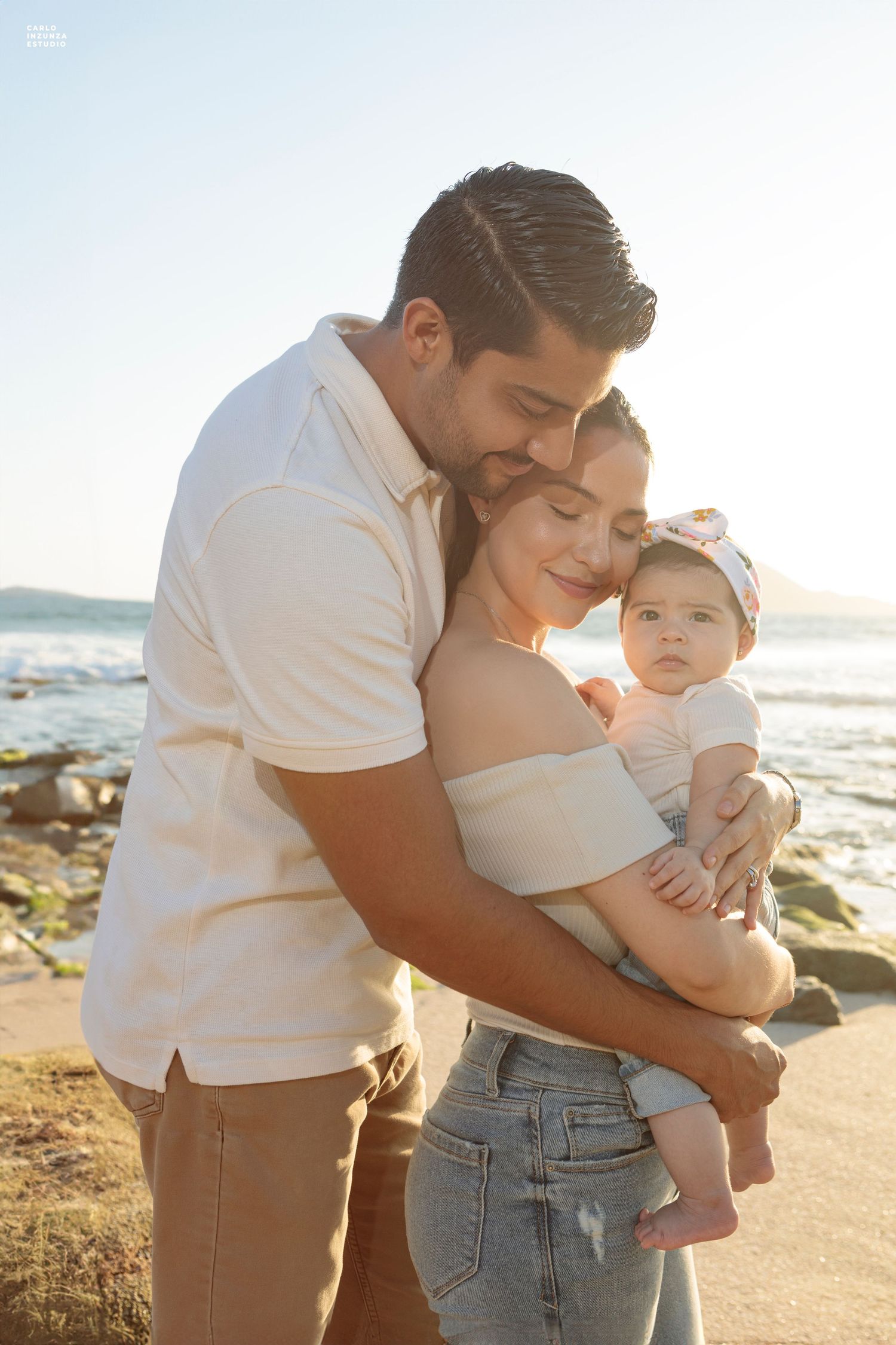 A loving couple tenderly embraces their baby girl on a sunlit rocky beach at golden hour.