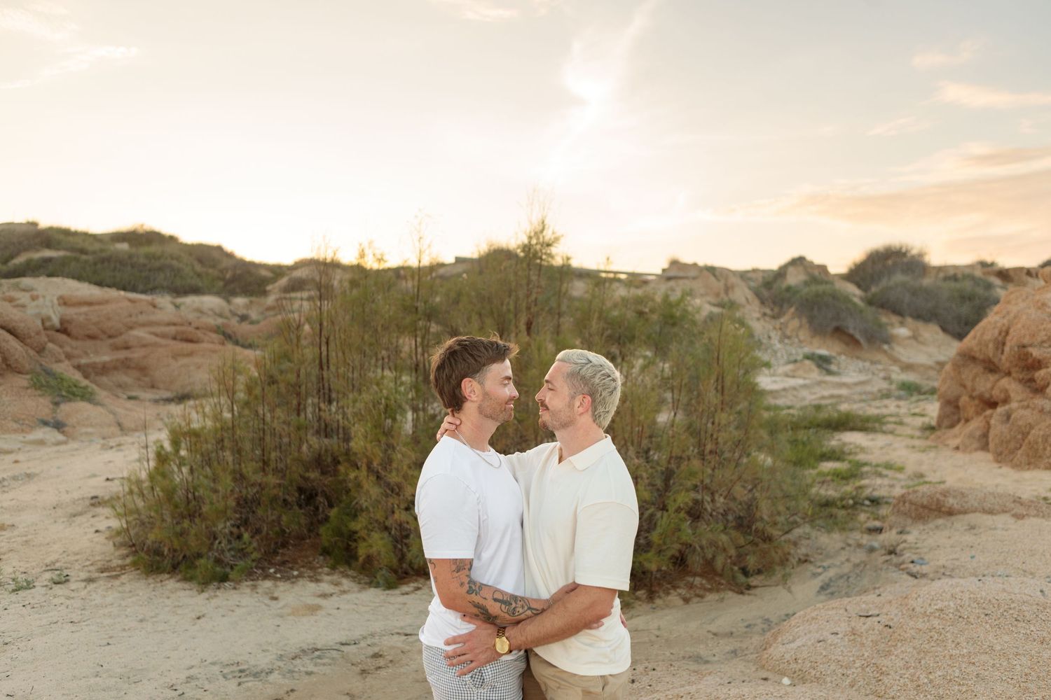 Two men holding each other and gazing into eyes at sunset in sandy desert landscape.