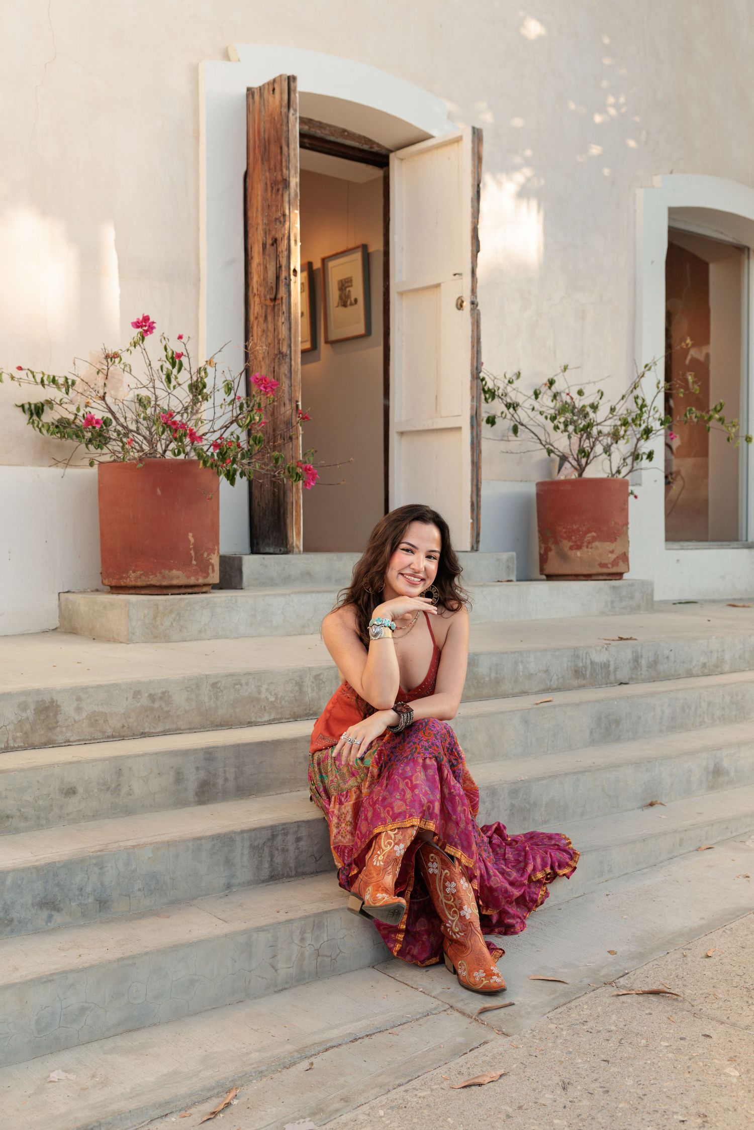 Woman in colorful pink and orange sari sitting on stone steps outside a white building with potted flowers.