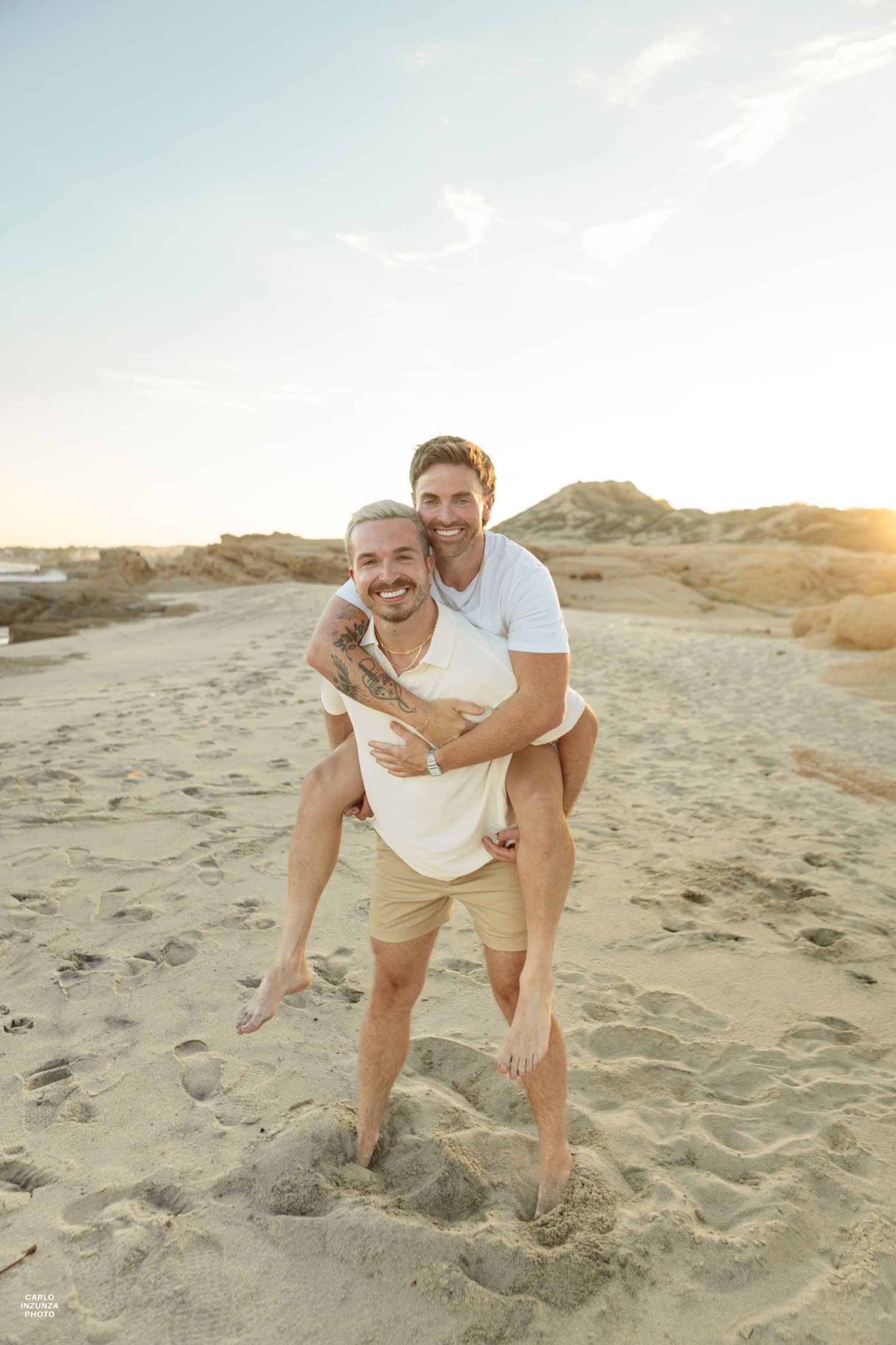 Man giving piggyback ride to partner on sandy beach while both smile at sunset.