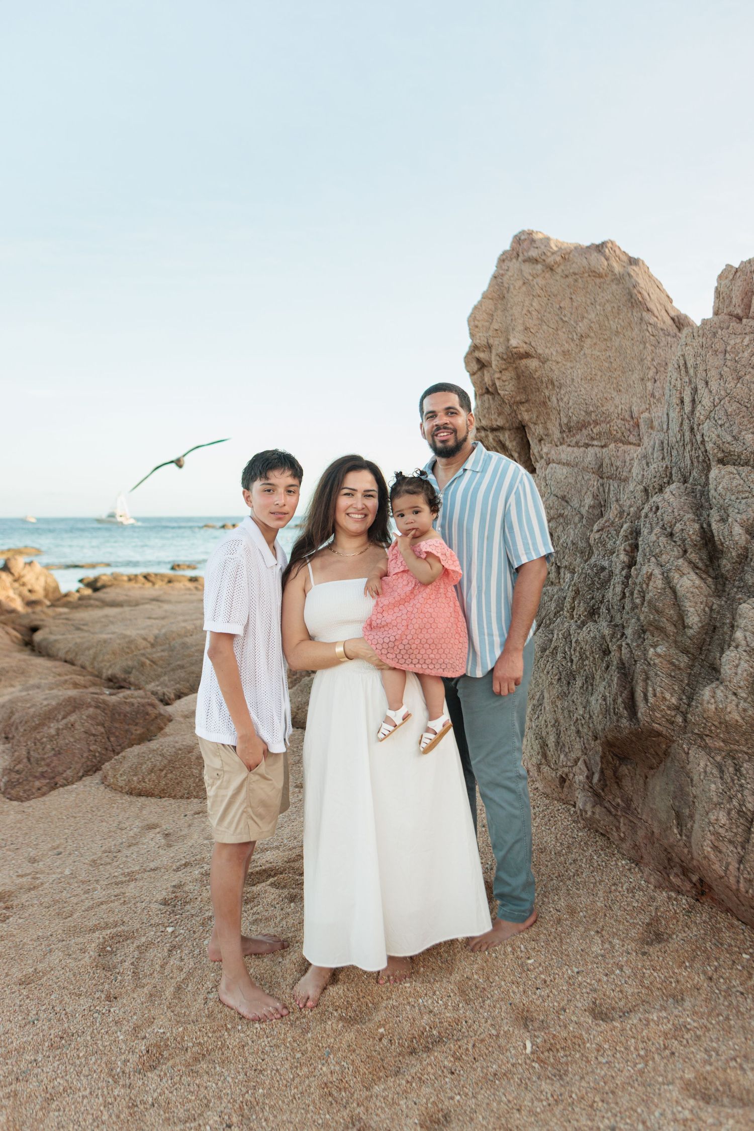 Family of four posing near rocky beach, parents holding toddler girl in pink dress, older son beside them.