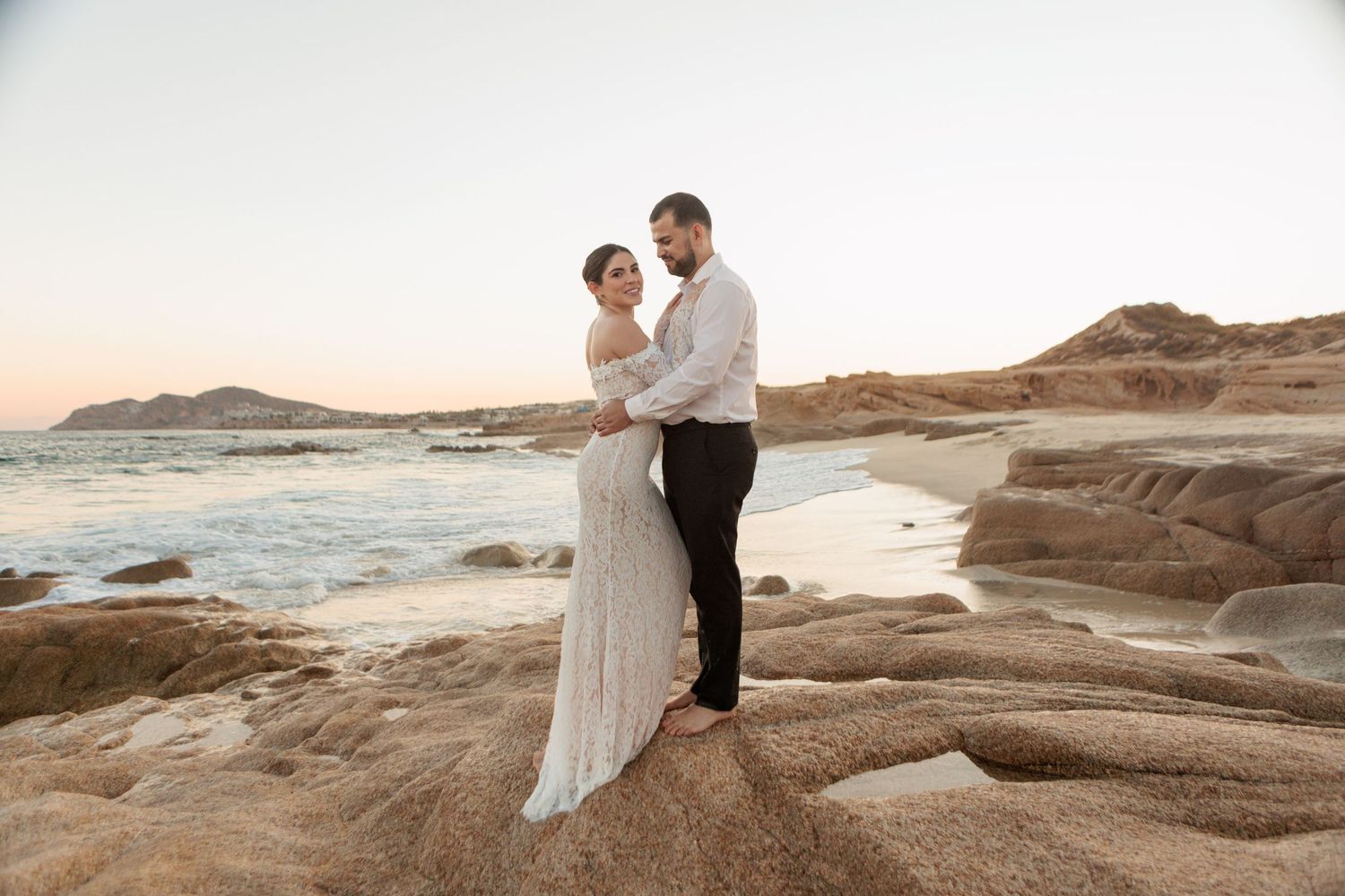 Bride and groom embrace on rocky seaside cliffs at golden hour during their beach wedding photoshoot.