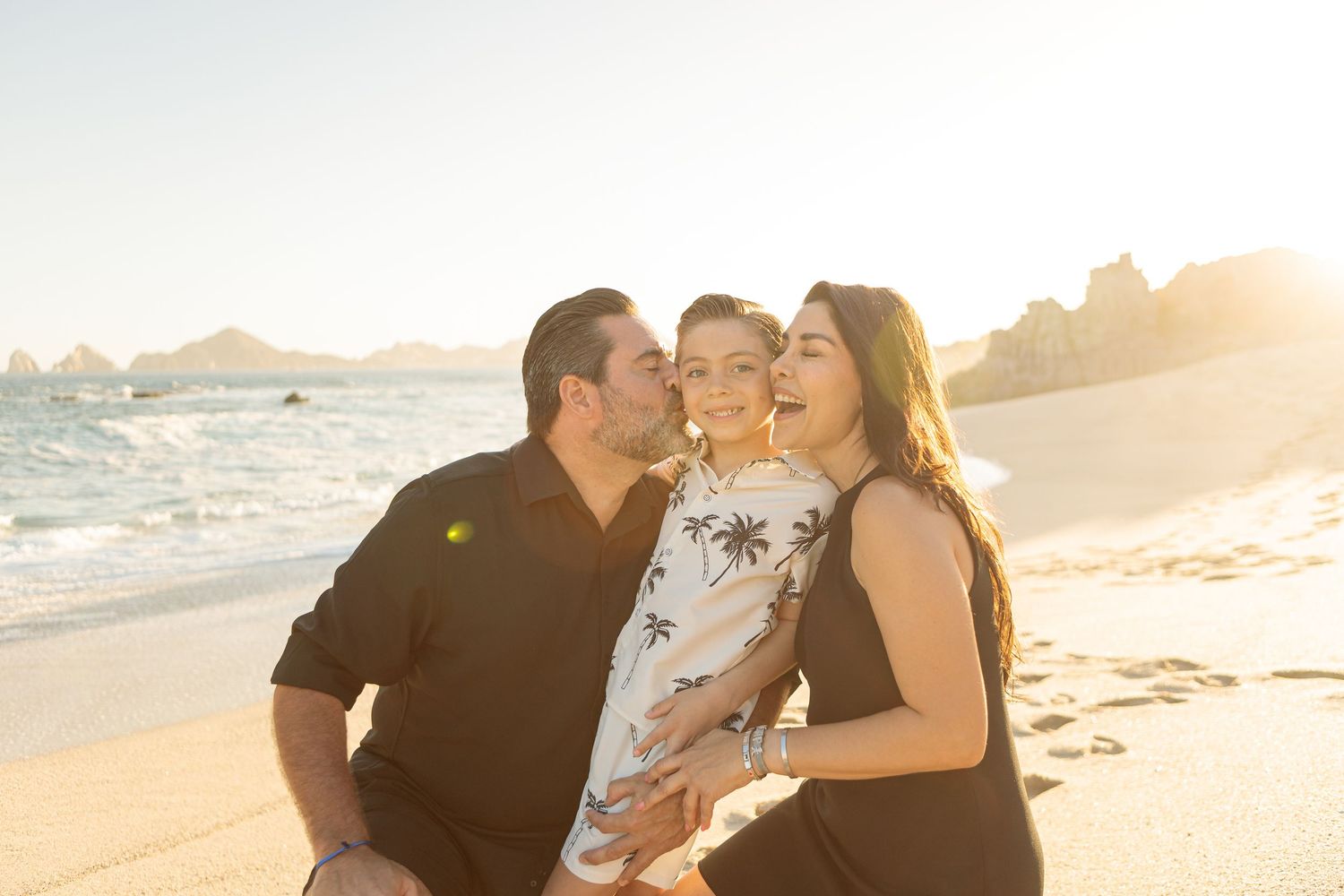 Family of three sharing a joyful moment on a sunny beach, parents kissing child on cheeks at golden hour.