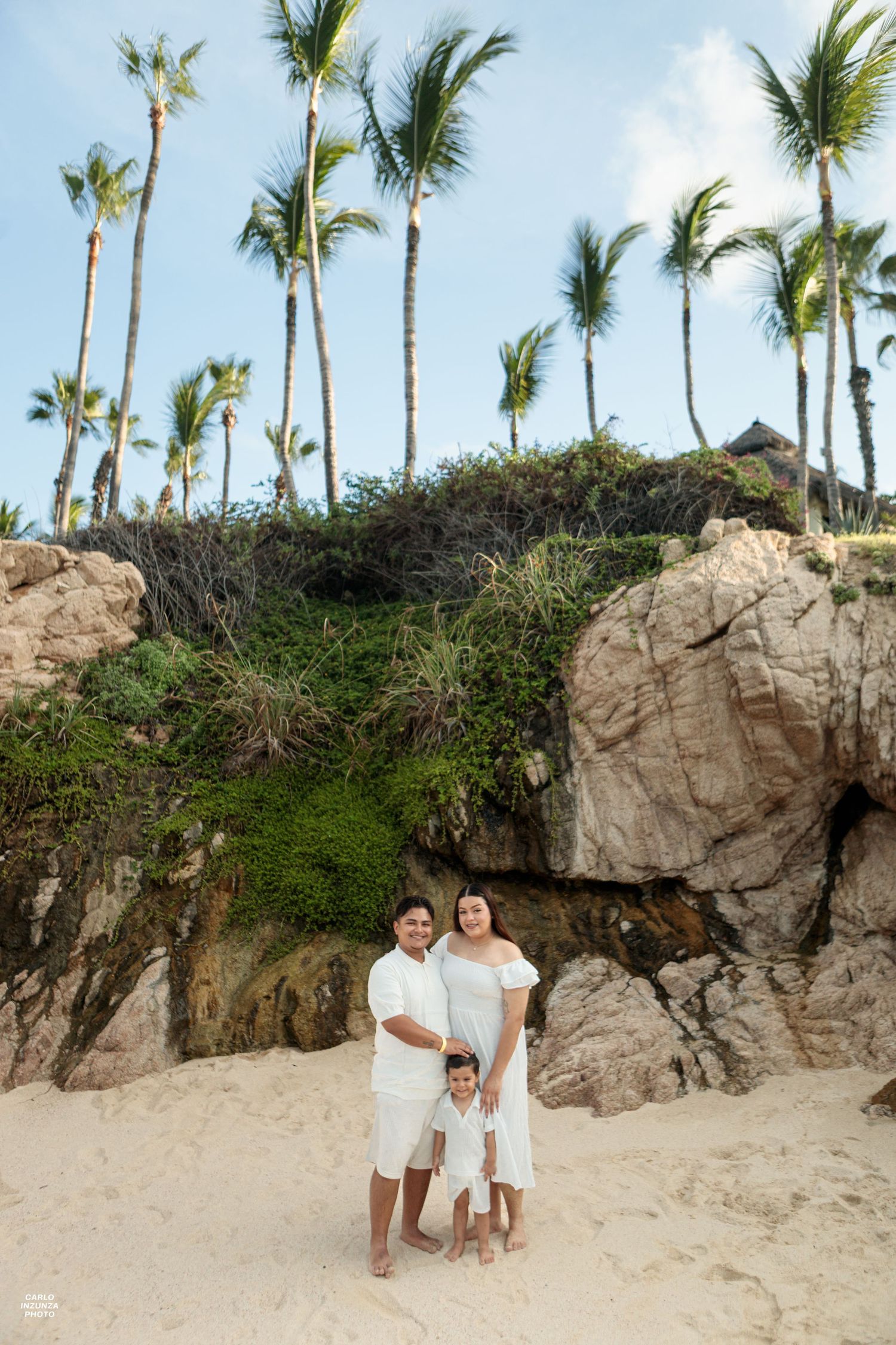 Family of three in white outfits stands on tropical beach with palm trees.
